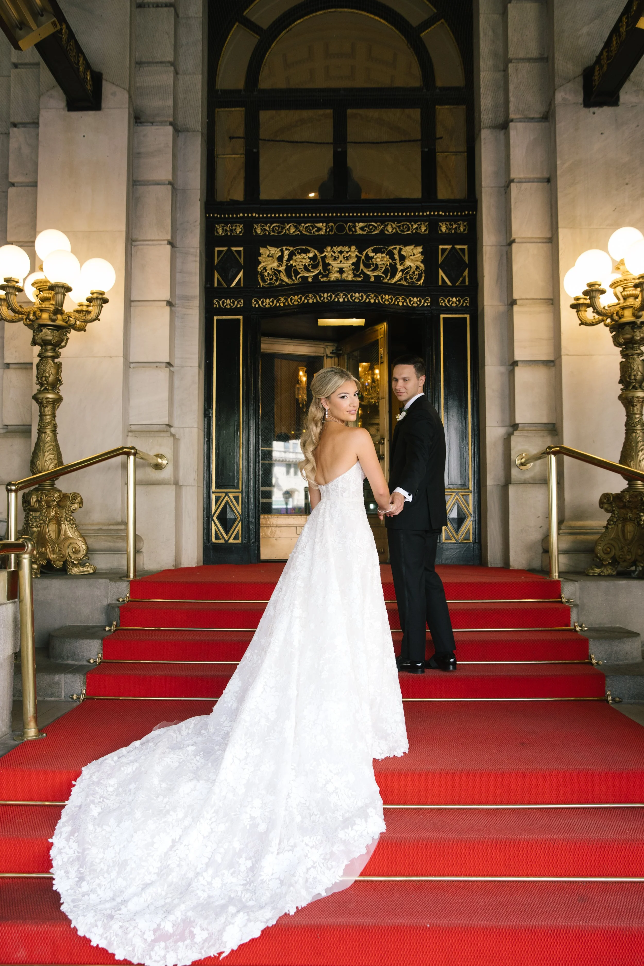 A bride and groom standing on red carpeted stairs outside a grand building, holding hands and looking at each other. The bride wears a white wedding gown with a long train, and the groom is dressed in a black tuxedo.