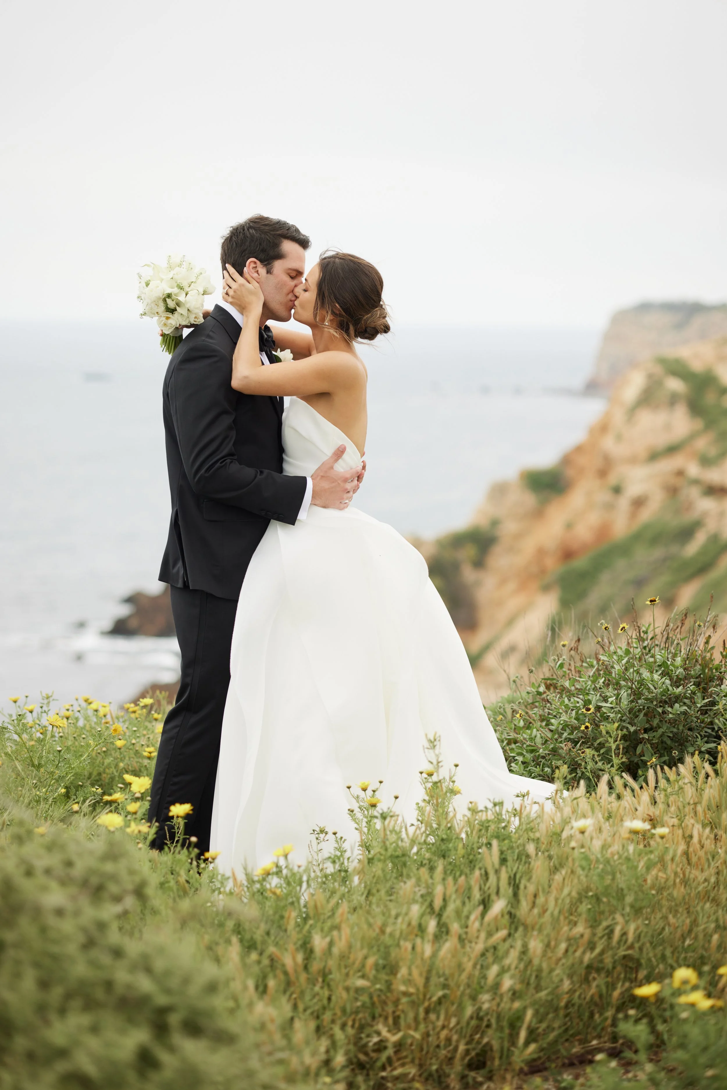 A bride and groom sharing a kiss outdoors on a grassy area with yellow flowers, overlooking a rocky coastline and ocean in the background.