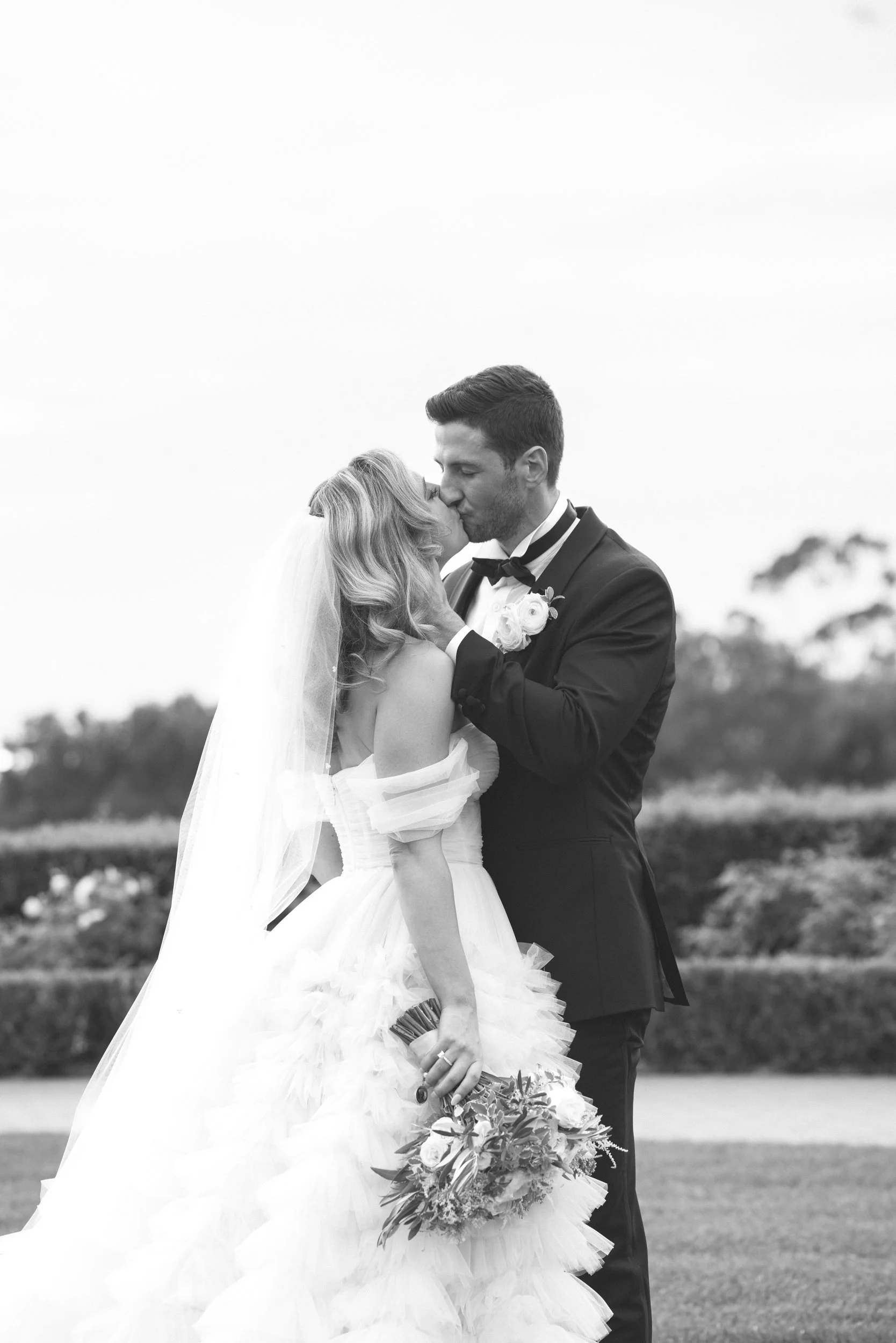 A wedding couple, the bride in a strapless white wedding gown with a layered skirt, holding a bouquet of flowers, and the groom in a tuxedo with a bow tie and boutonniere, embracing and kissing outdoors with blurred trees in the background.