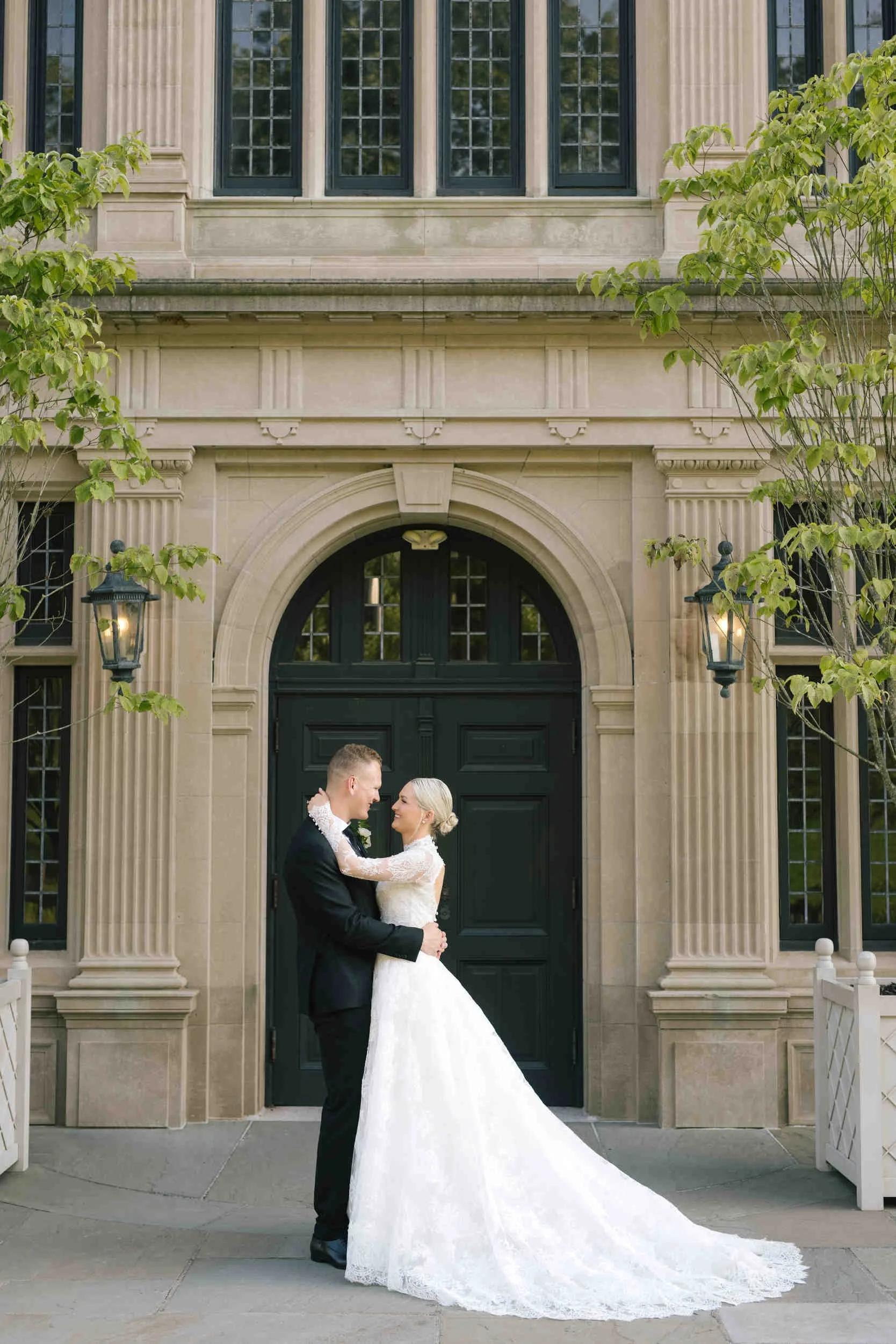 A bride and groom standing in front of a large building with columns and black doors, holding each other and smiling.