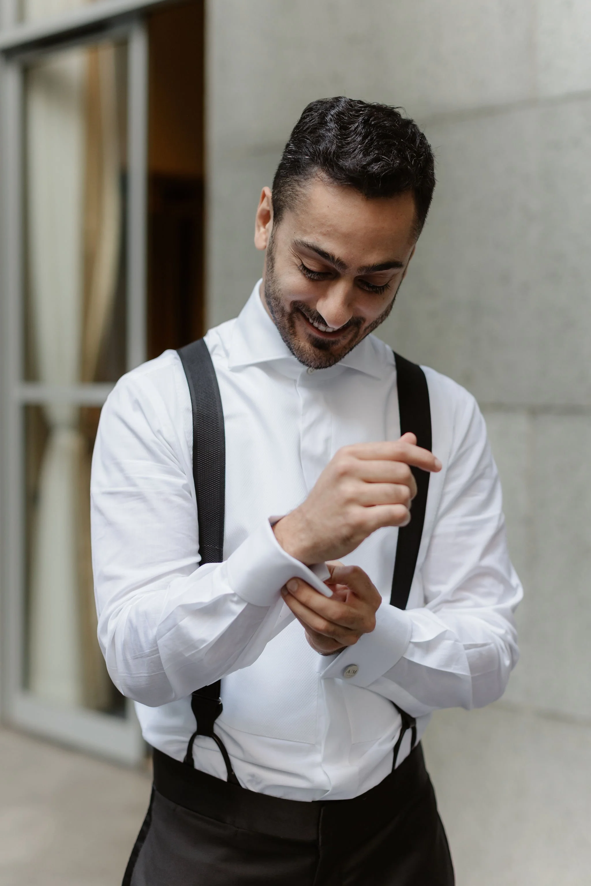 A man with dark hair and a beard, dressed in a white dress shirt with black suspenders, smiling while looking down and adjusting something on his shirt in a hallway with a gray wall.