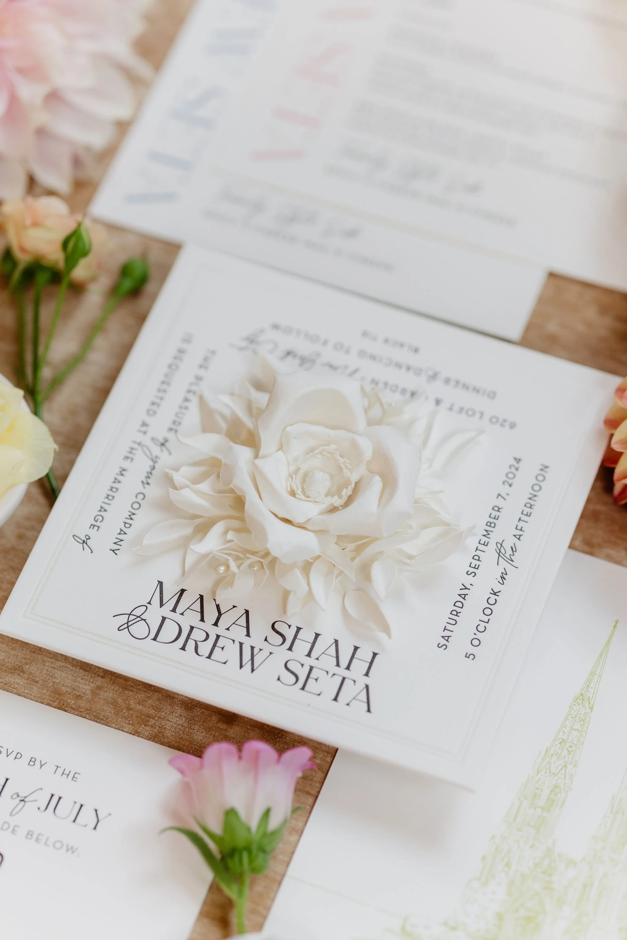 Close-up of wedding invitation card with white floral decoration, surrounded by light pink and peach flowers, on a wooden surface.