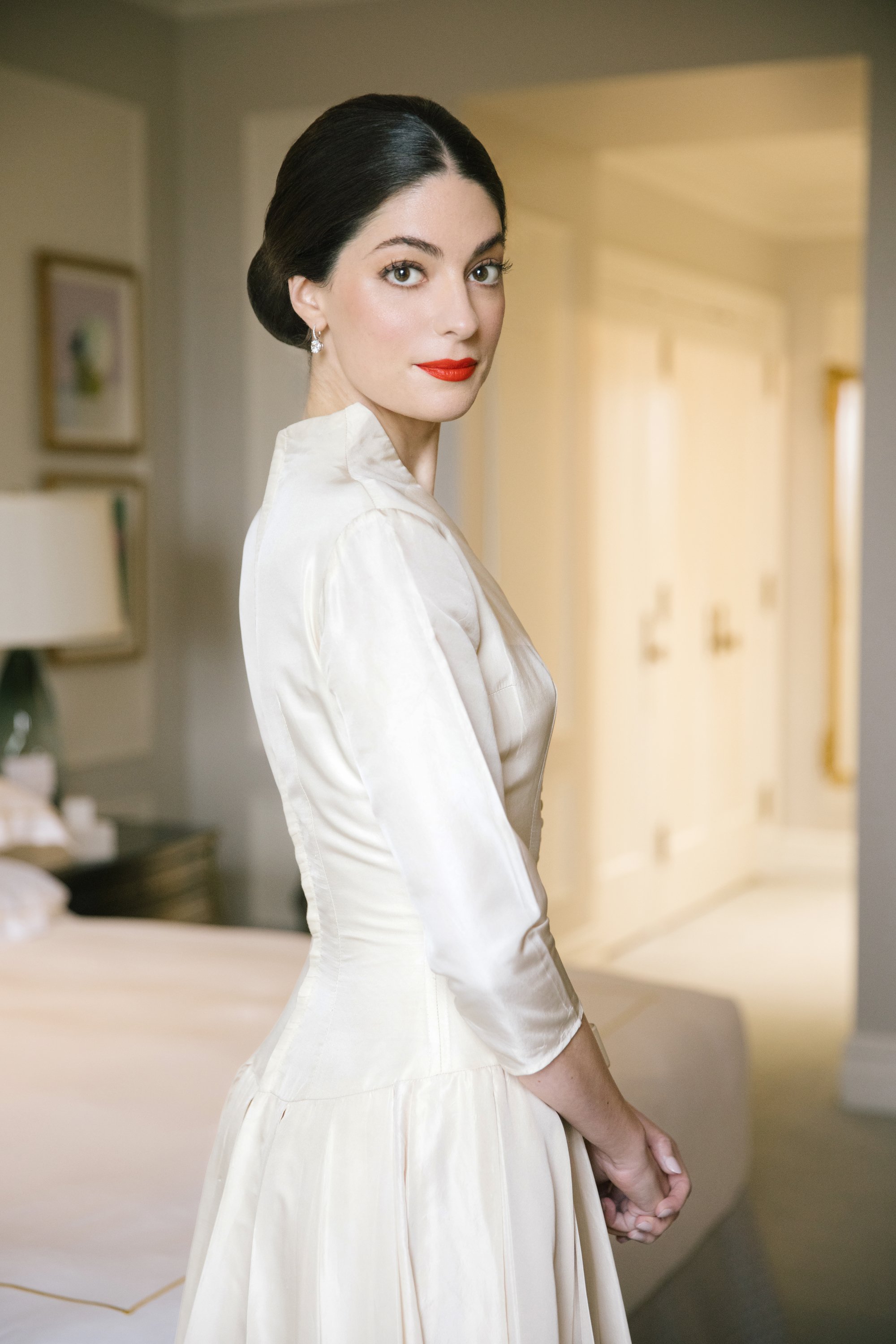 A woman with dark hair styled in a vintage updo, wearing pearl earrings and red lipstick, dressed in a white satin dress, standing indoors with a neutral background.