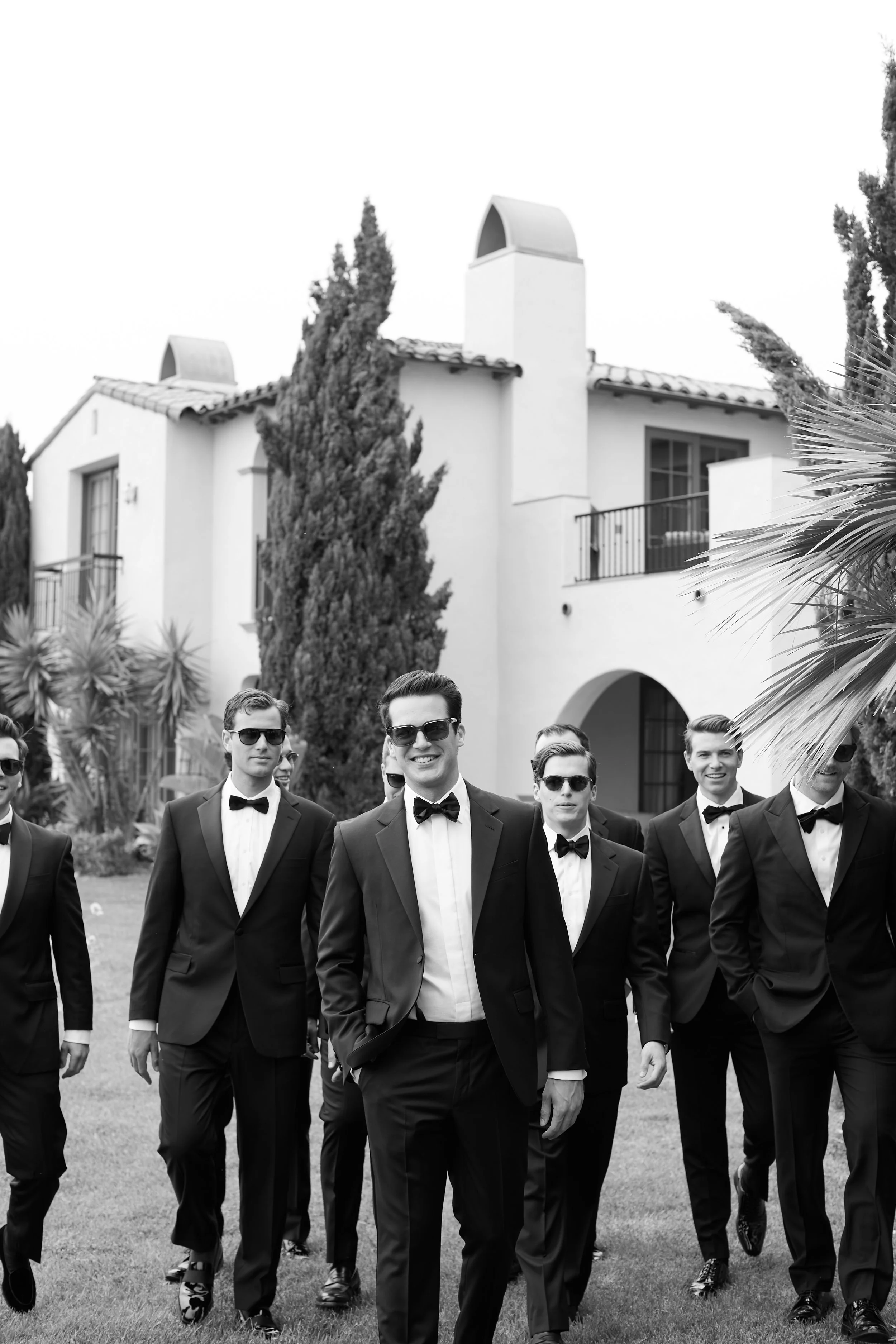 A group of men dressed in formal tuxedos walking outdoors on a lawn in front of a large white house with trees and plants.