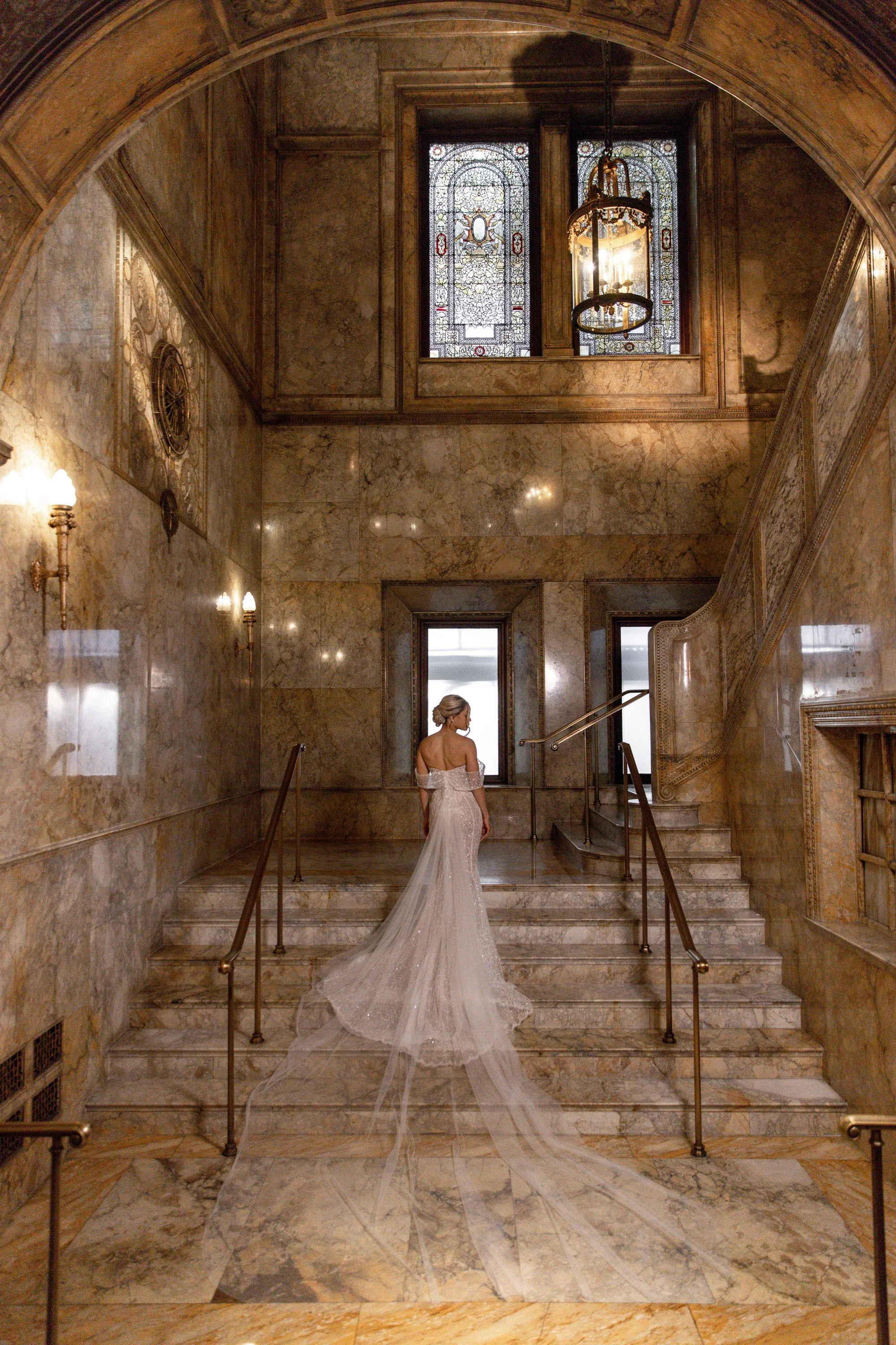 A woman in a white wedding dress with a long train standing on marble stairs inside a grand building with marble walls, stained glass windows, ornate lighting fixtures, and a curved staircase railing.