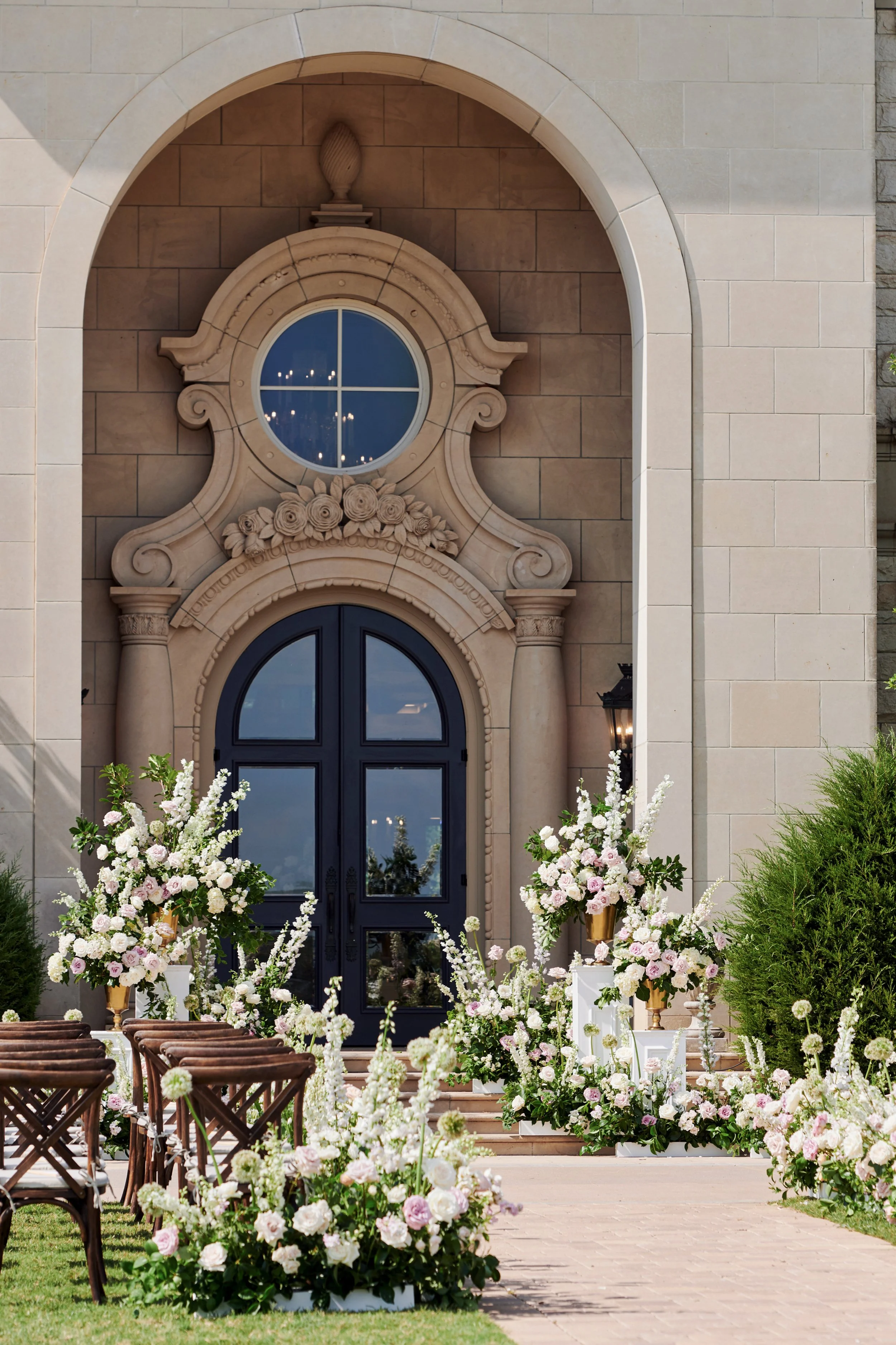 Entrance of a building with a black arched door, decorated with floral arrangements including white and pink flowers, and greenery, indicating a wedding or special event setting.