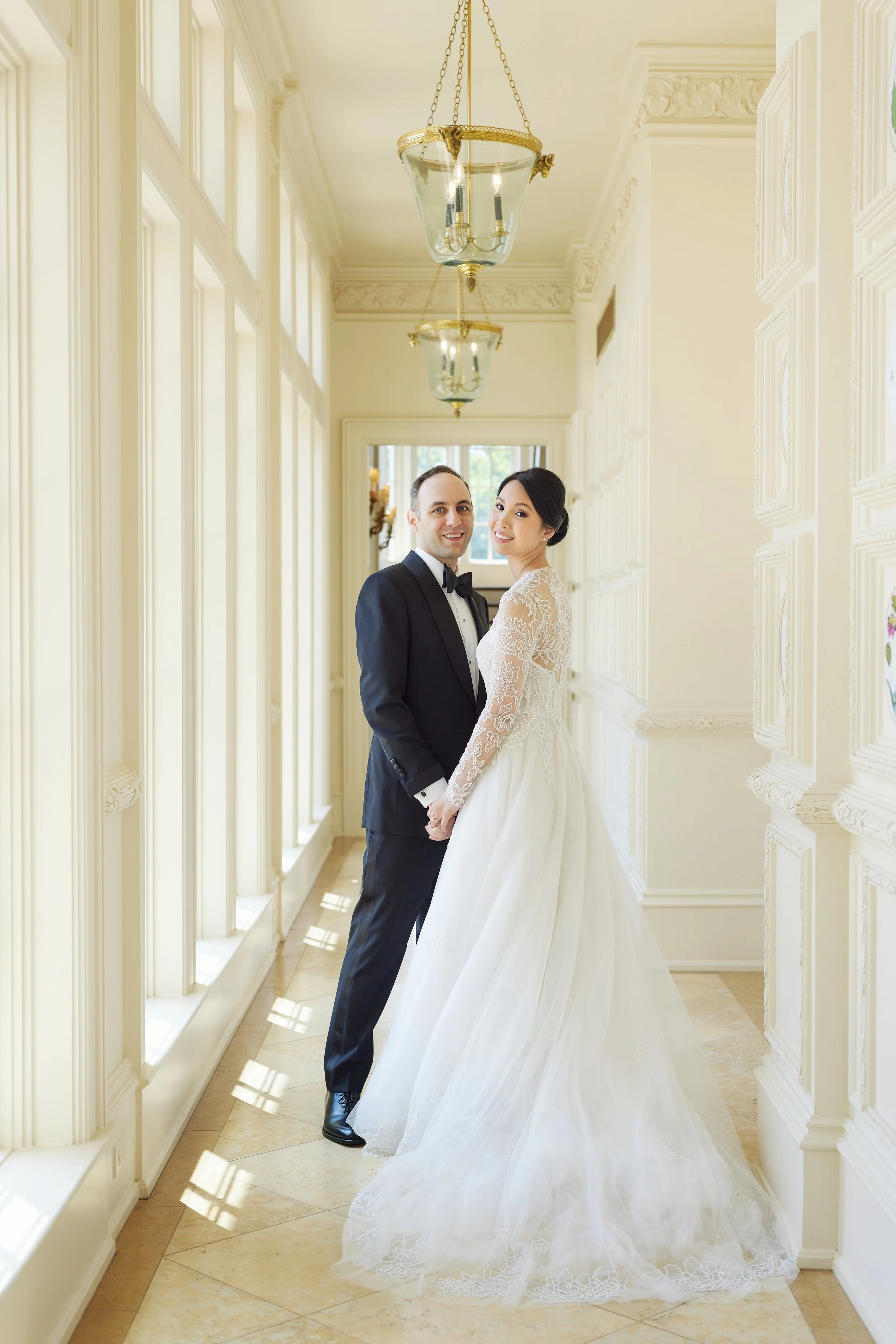 A newlywed couple holding hands and smiling in a bright, elegant hallway with large windows and ornate ceiling details. The bride wears a lace wedding gown with a long train, and the groom is dressed in a tuxedo.
