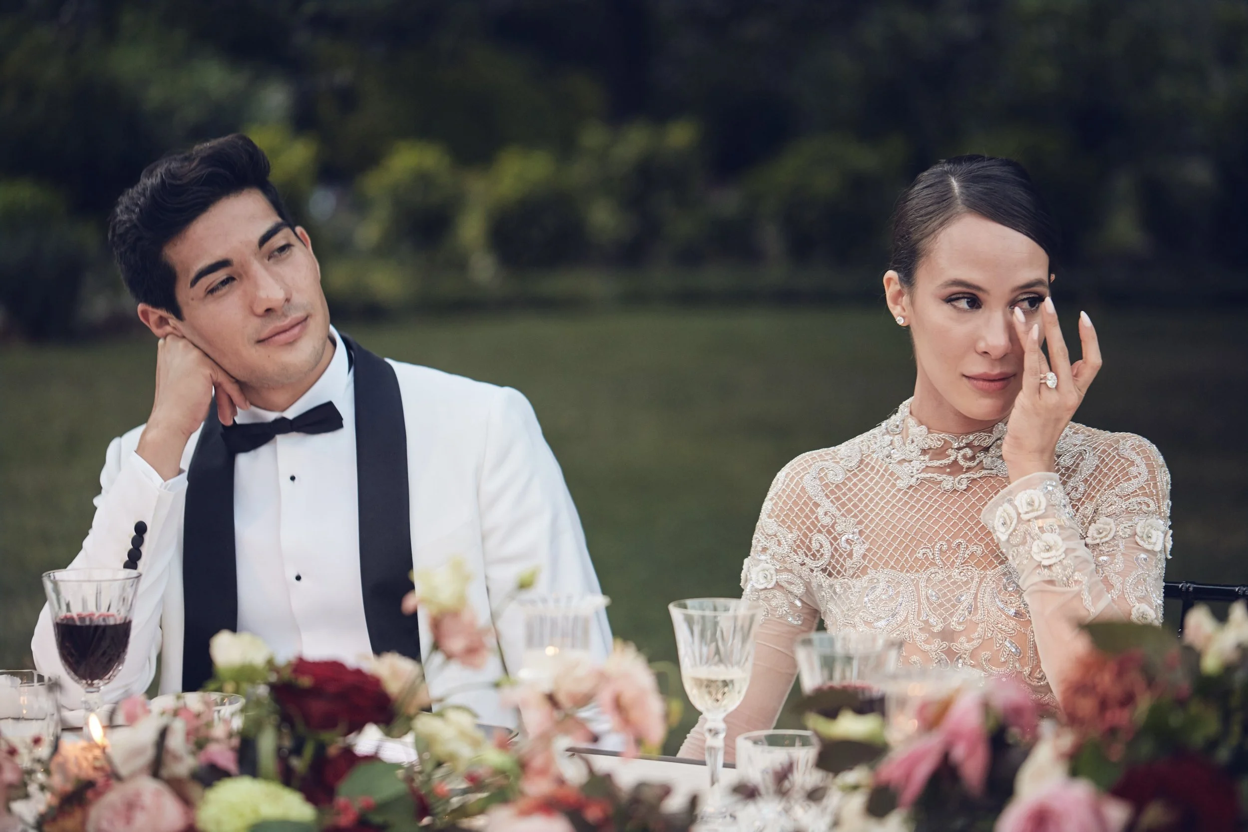 A man in a tuxedo and a woman in an elegant lace dress sit at a dining table outdoors during a formal event, with flower arrangements and glasses of wine on the table.