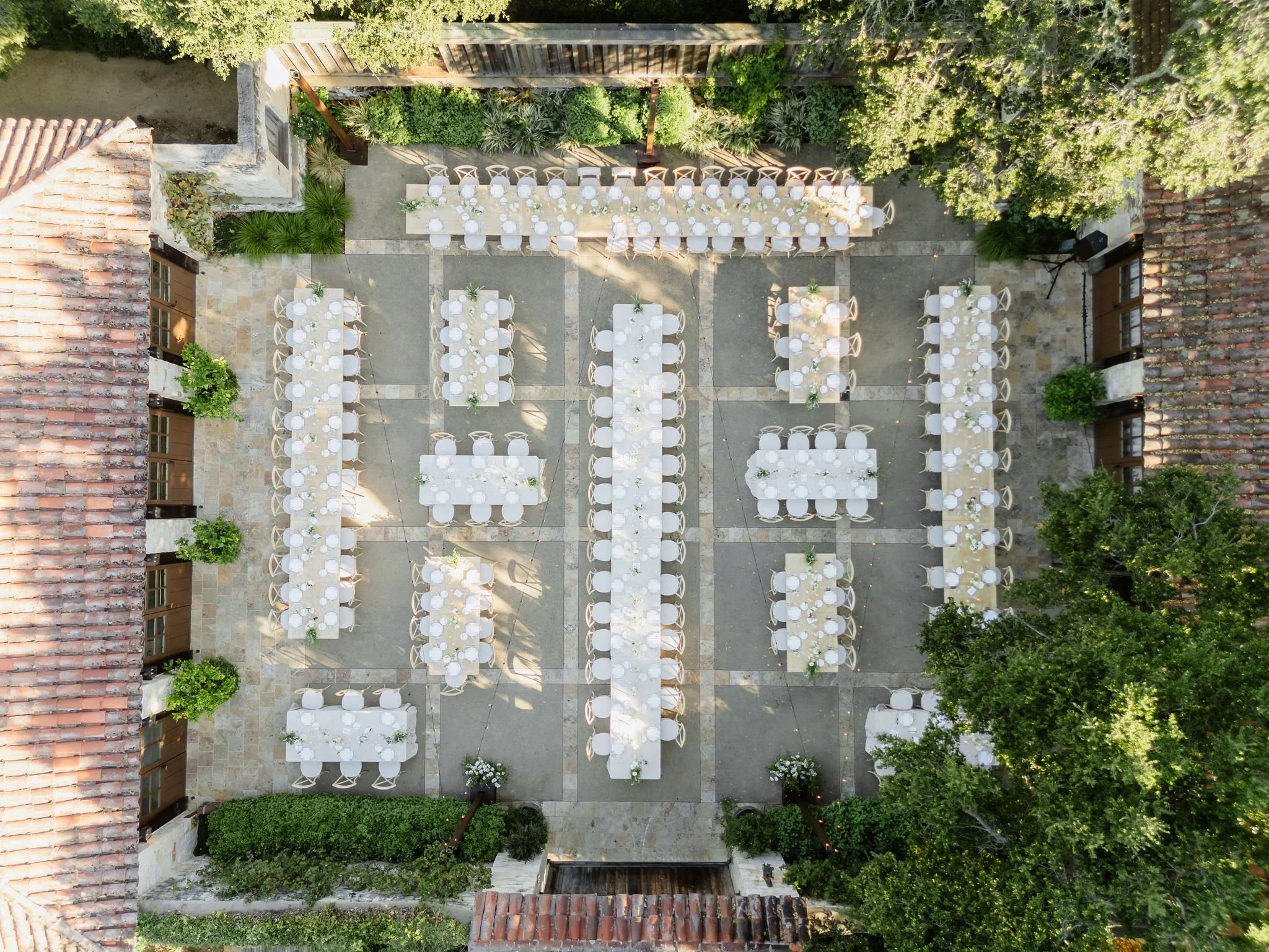 Outdoor event setup with long banquet tables, round tables, and chairs under trees, surrounded by buildings with tiled roofs.
