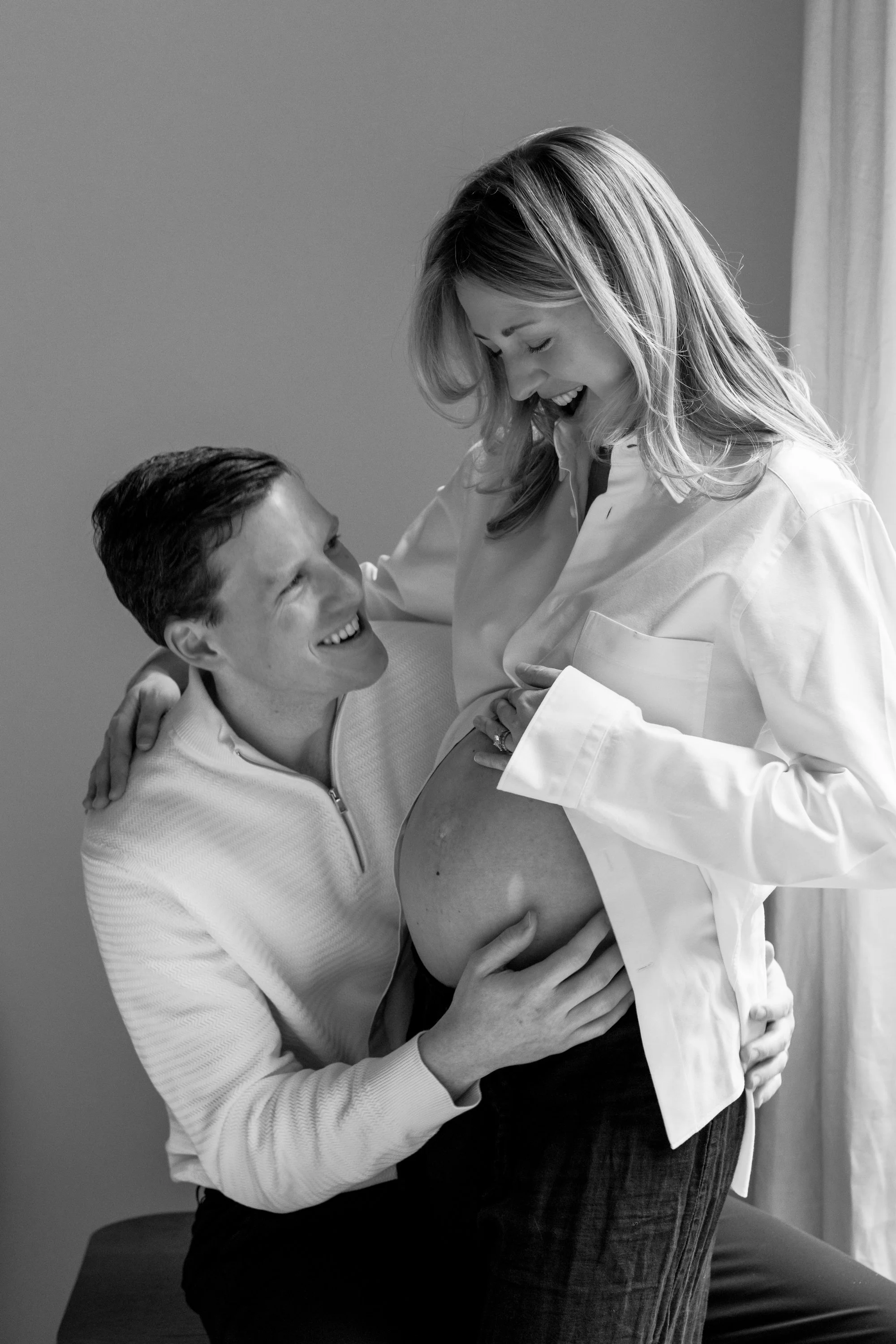 A couple, a man and a woman, smiling and enjoying a moment together. The woman is pregnant and cradling her belly while the man is hugging her and looking at her affectionately.