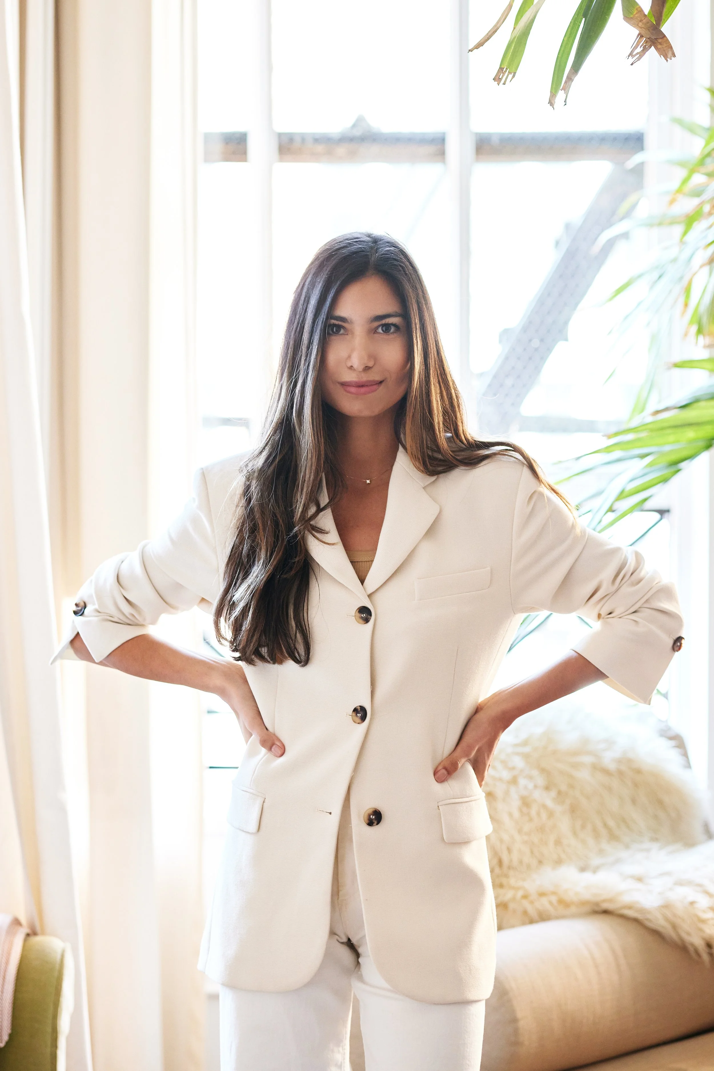 A woman with long dark hair wearing a white blazer and matching pants standing indoors with her hands on her hips in front of large windows and green plants, with a beige couch and fluffy throw blanket visible in the background.