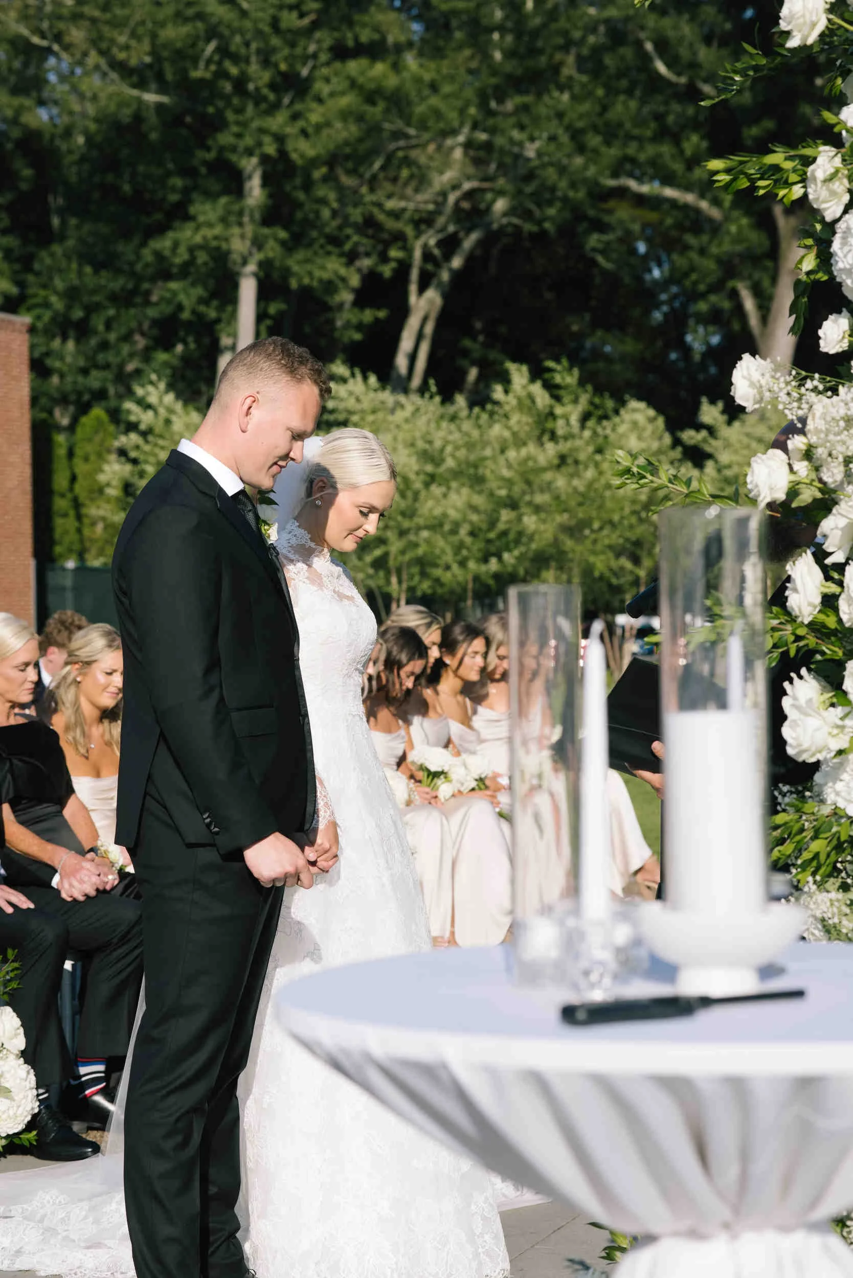 A bride and groom kneeling with heads bowed during a wedding ceremony outdoors, surrounded by seated guests and decorated with white flowers.