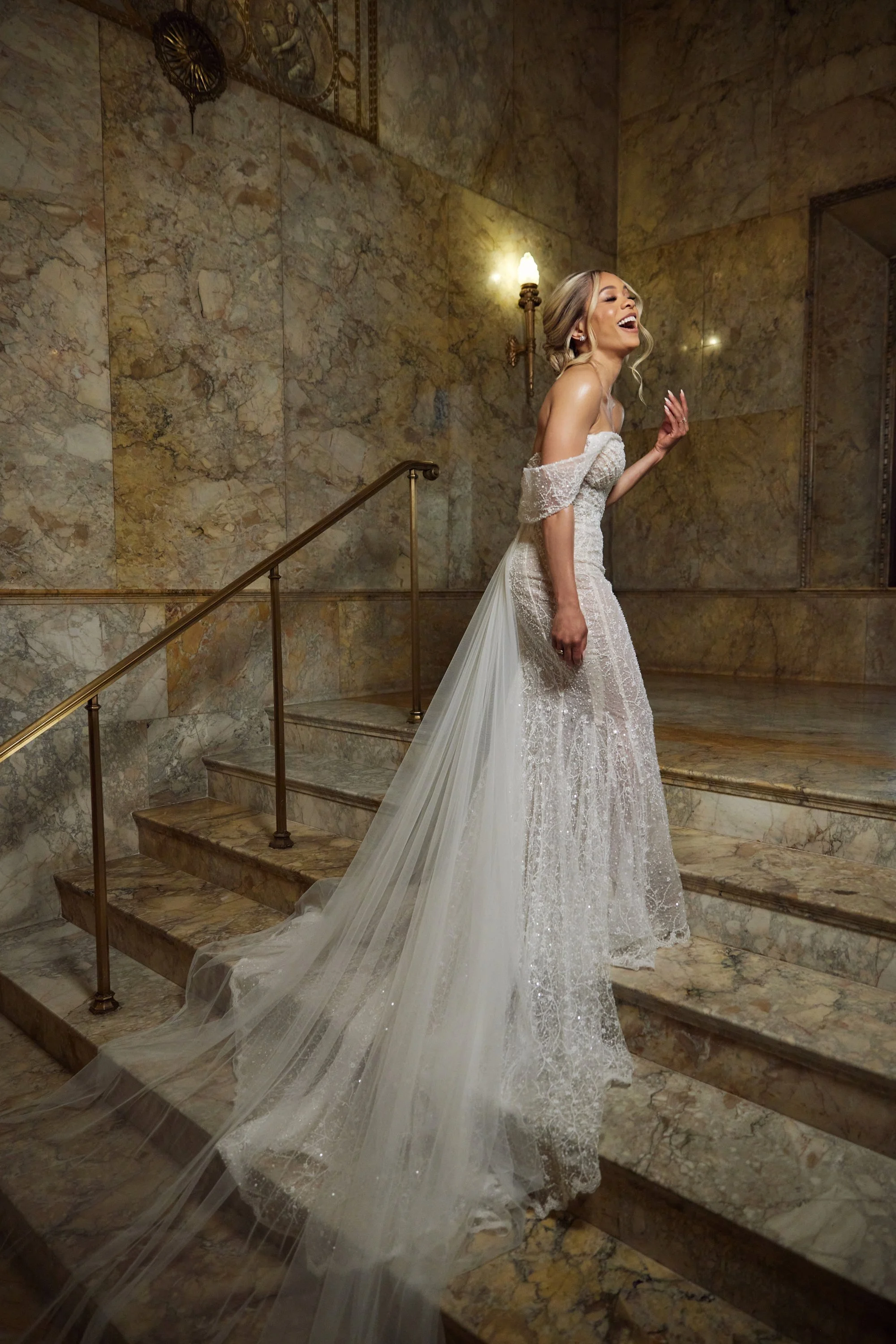 A woman in a wedding dress standing on marble stairs, laughing with her eyes closed inside a marble-paneled room with warm lighting.