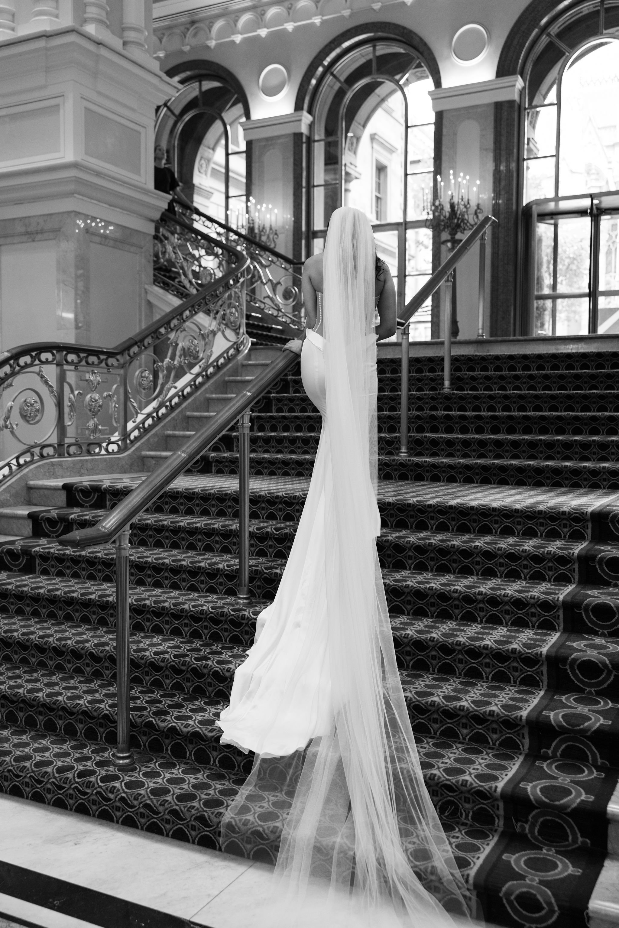 A woman in a long white gown and veil walking up a grand staircase inside a luxurious building with large arched windows and ornate chandeliers.