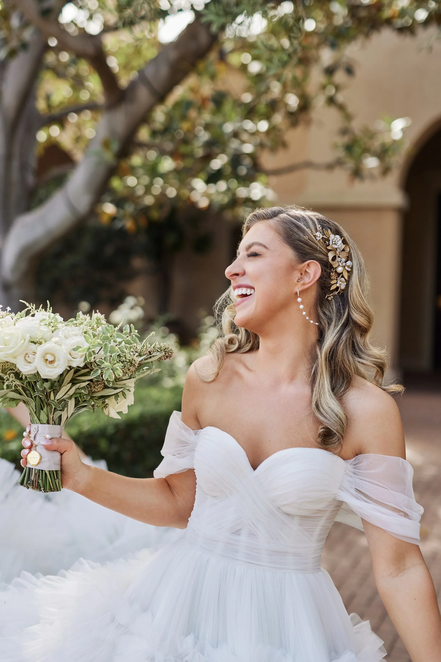 A smiling bride in a white wedding dress holding a bouquet of white and green flowers outdoors with trees and architecture in the background.