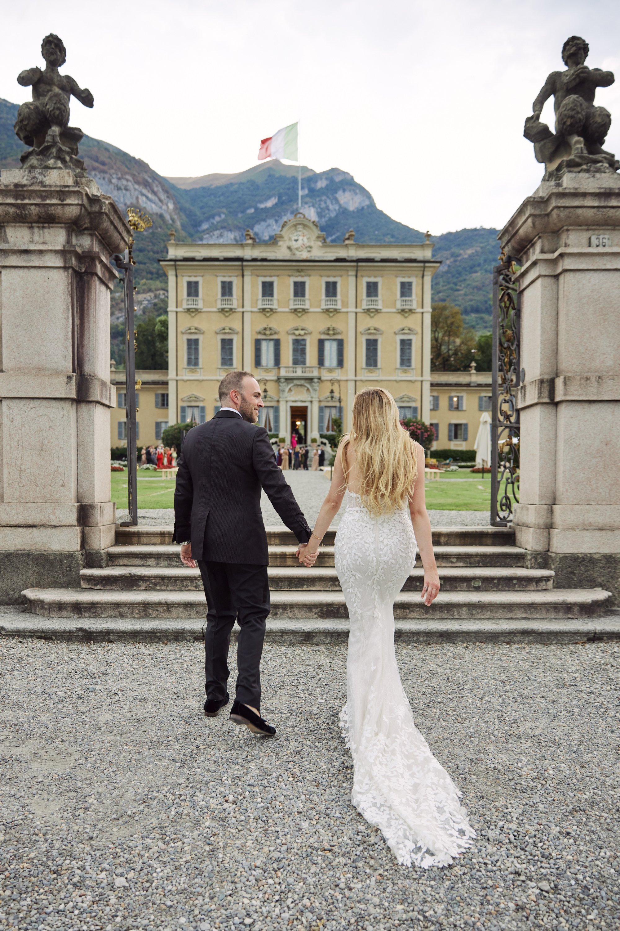 A bride and groom walking hand in hand on wedding day, in front of a large, elegant building with a flag on top, set against a mountainous backdrop.