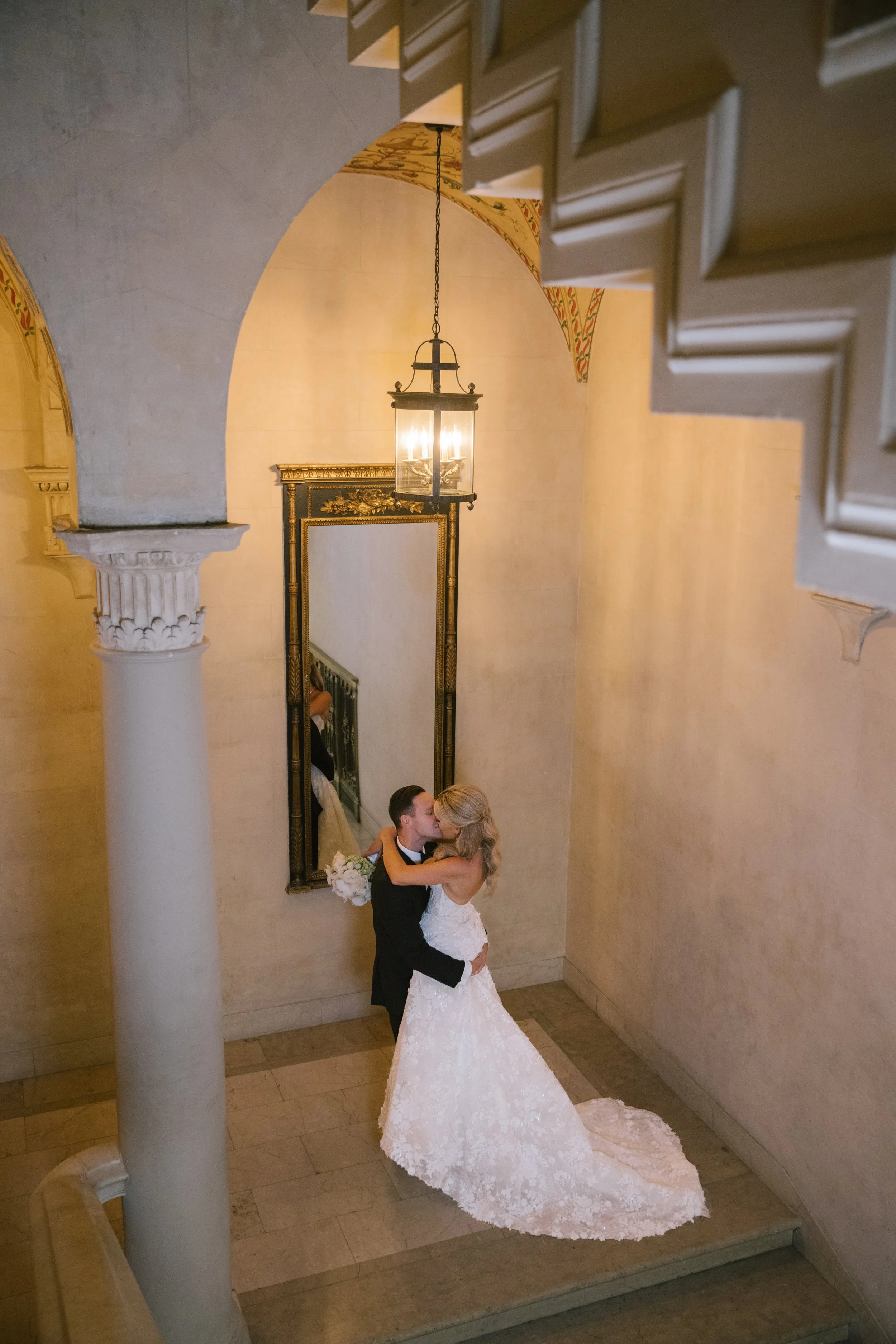 A bride and groom sharing a kiss in an elegant indoor space with arched ceilings, a vintage chandelier, and ornate details.