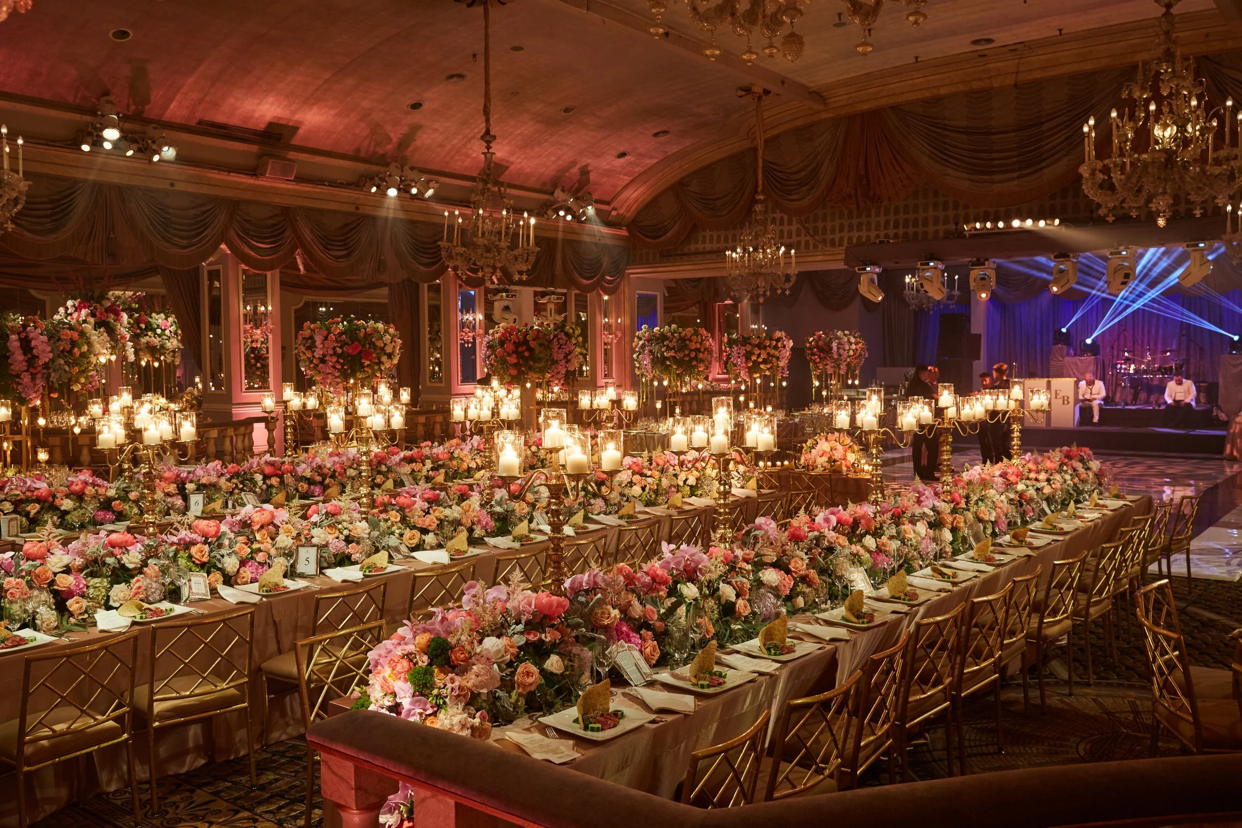 An elegantly decorated banquet hall with a stage and a microphone, filled with multiple round tables with floral centerpieces, candles, and chairs for guests, set for a formal event or celebration.