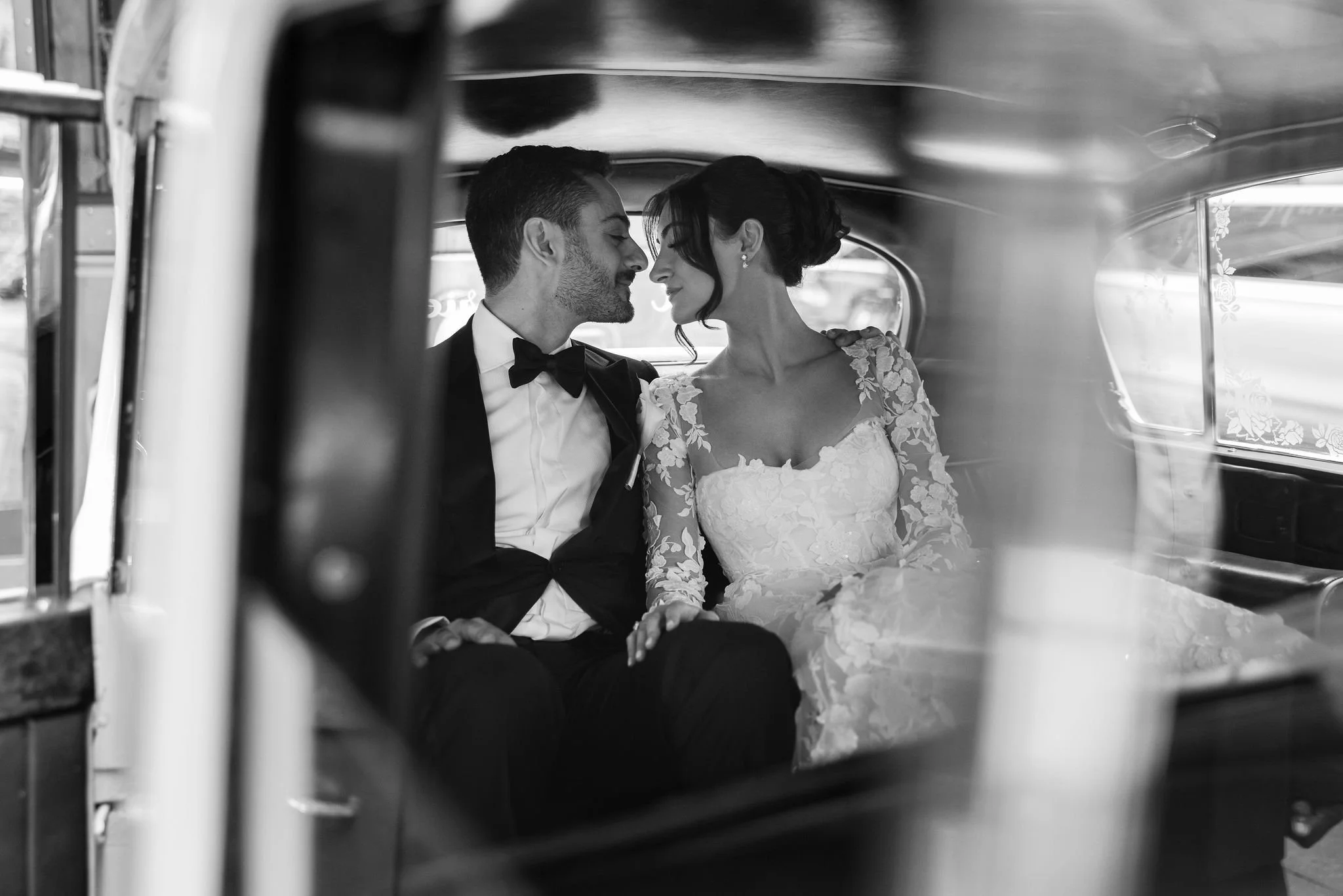 Black and white photo of a bride and groom sitting closely together inside a vintage vehicle, likely a car, with their foreheads touching and eyes closed, sharing an intimate moment.