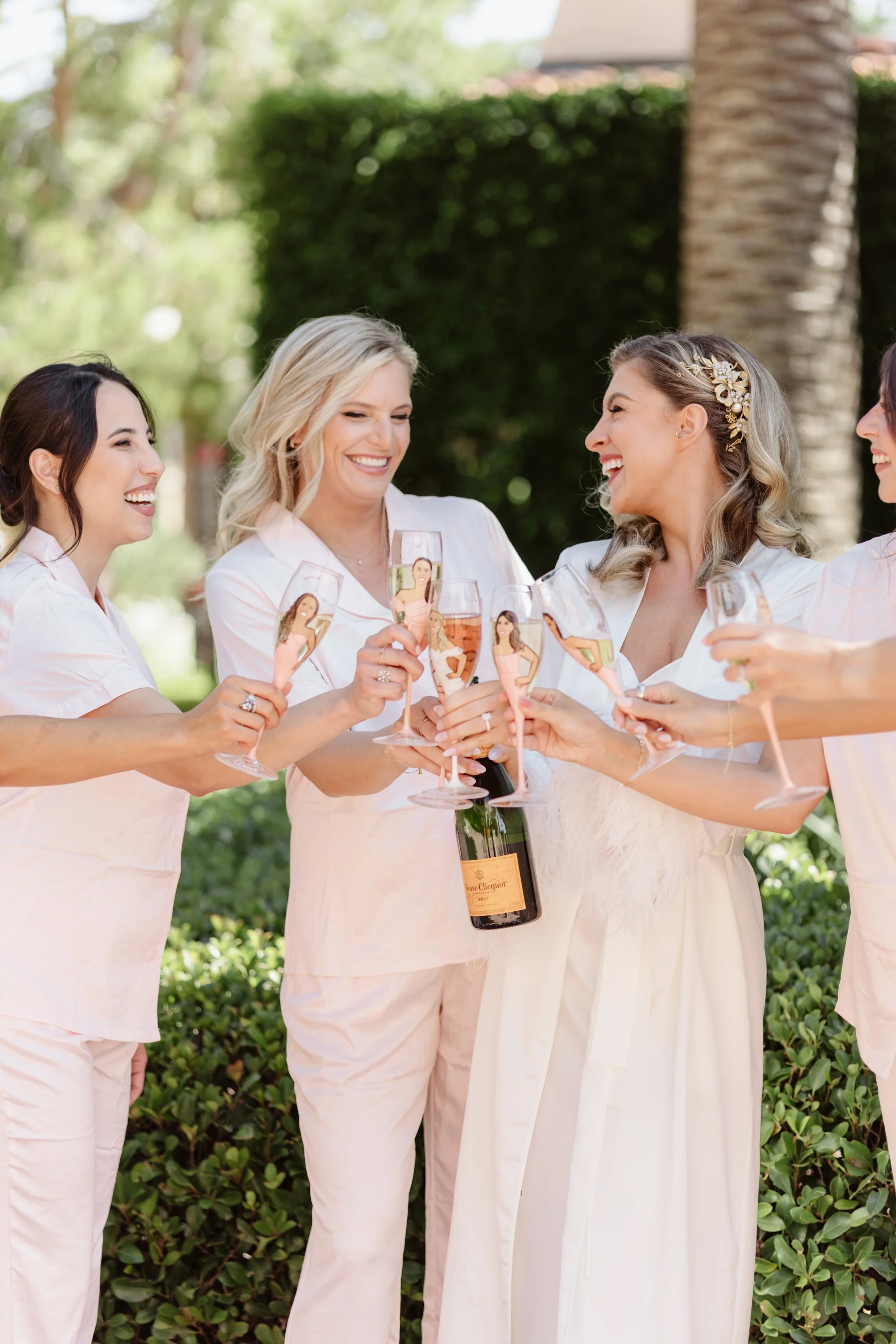 Group of women in cream-colored pajamas holding glasses of champagne and celebrating outdoors with a bottle of Veuve Clicquot in the center, smiling and laughing.