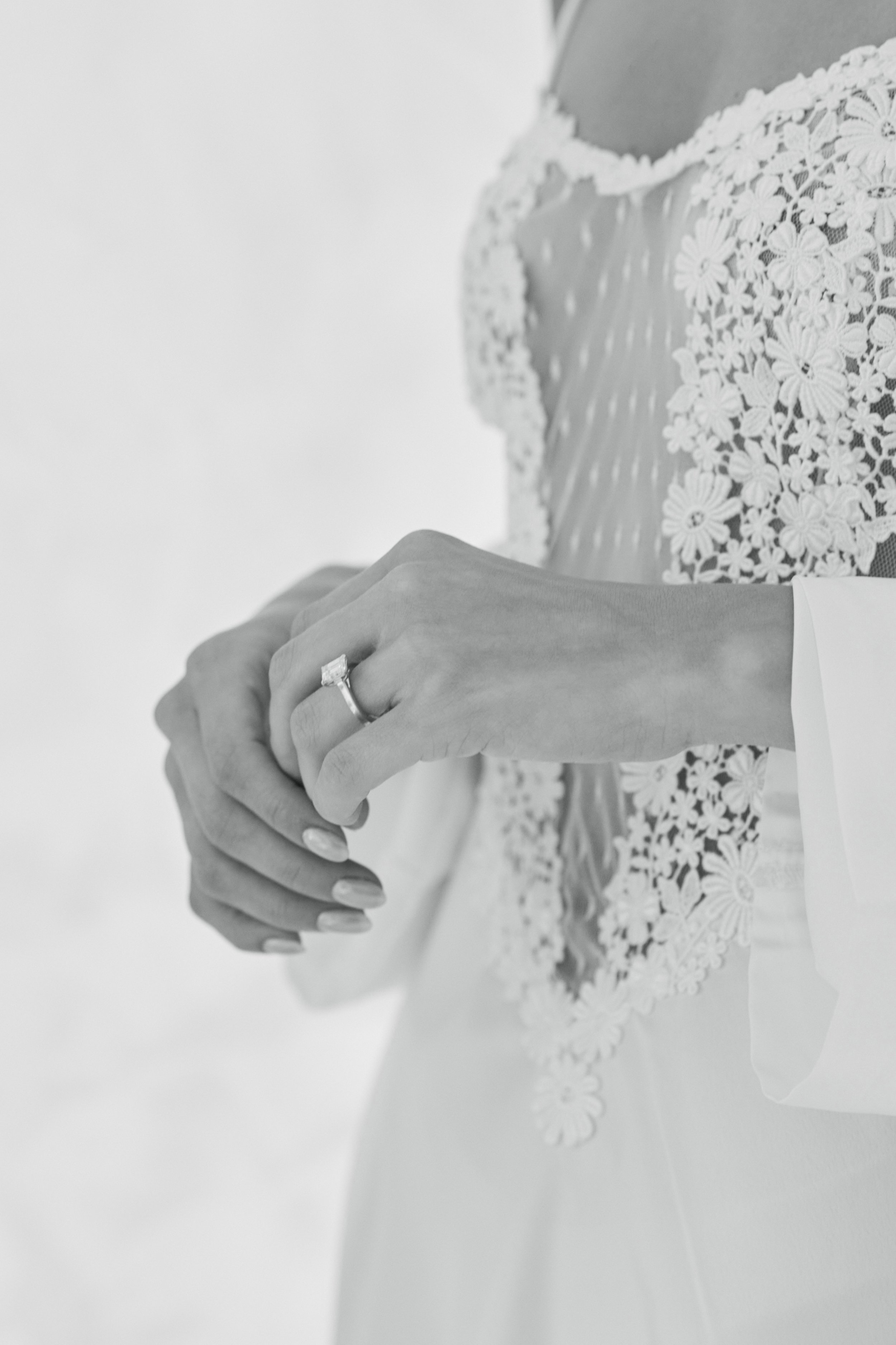 Close-up of hands clasped together, wearing a wedding ring, with lace fabric details on a wedding dress.