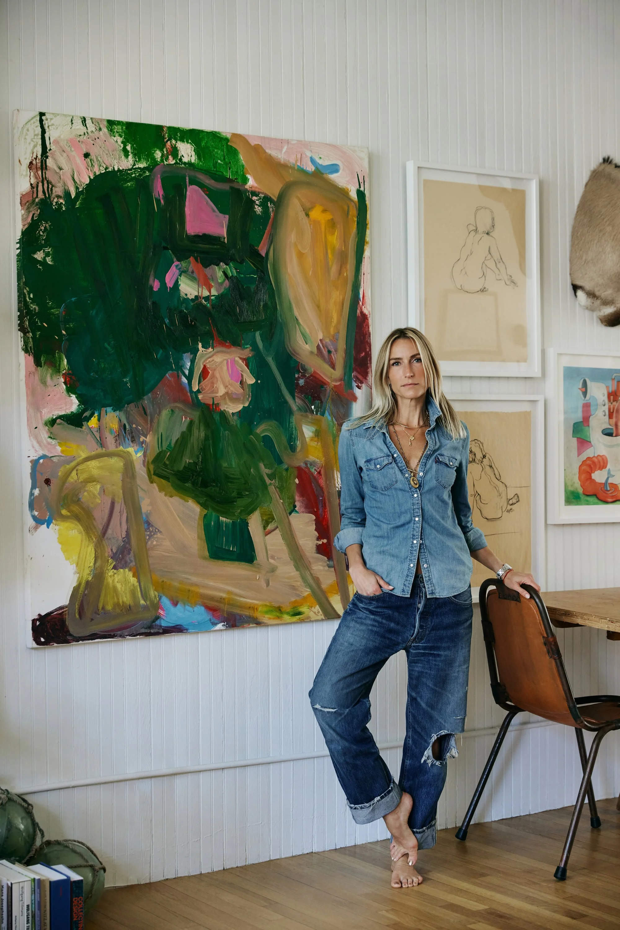 A woman in a denim shirt and distressed jeans standing barefoot in an art gallery, with abstract paintings on the white wall behind her and a chair nearby.