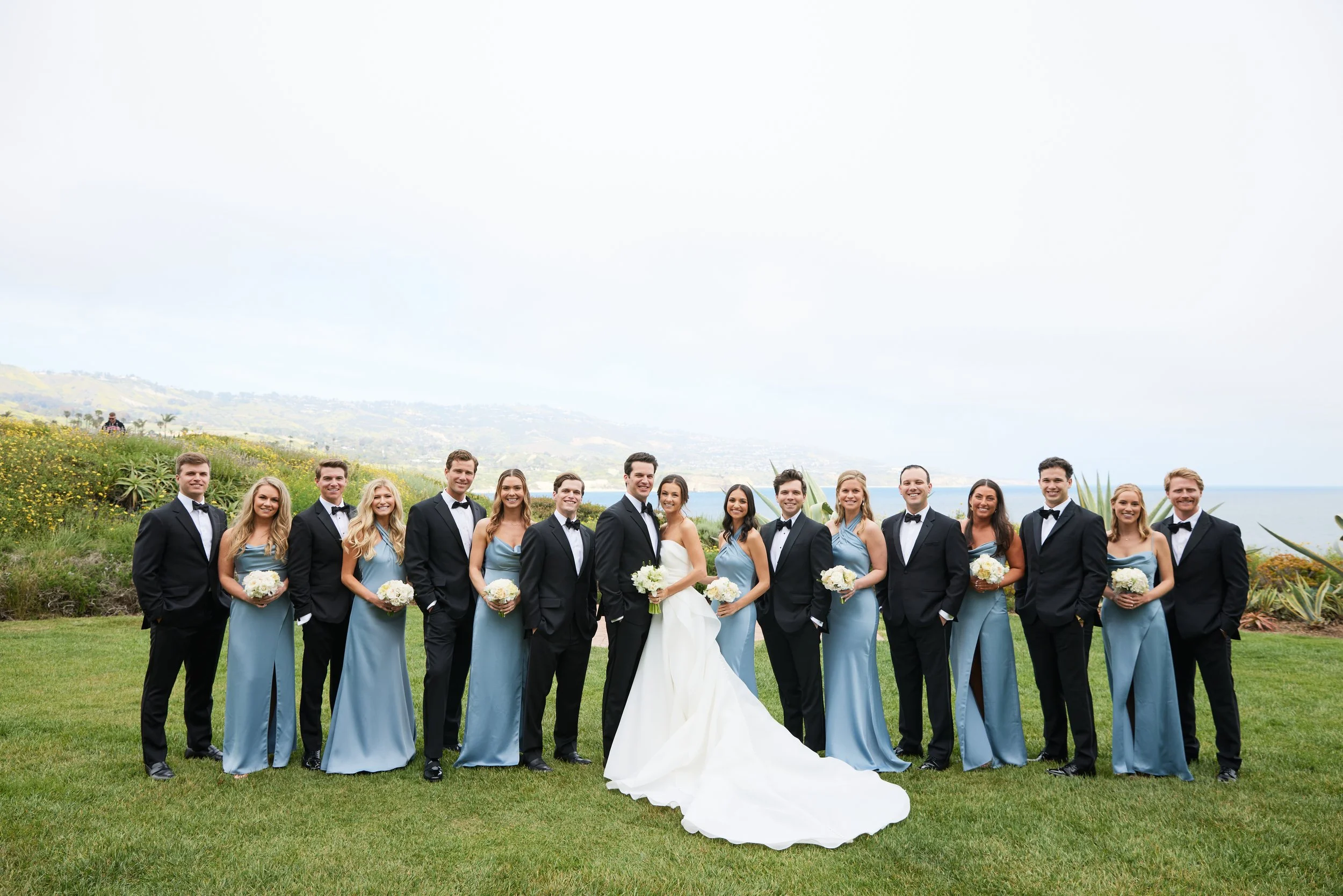 Group of people in formal wedding attire standing outdoors on a grassy area with a scenic view of hills and water in the background.