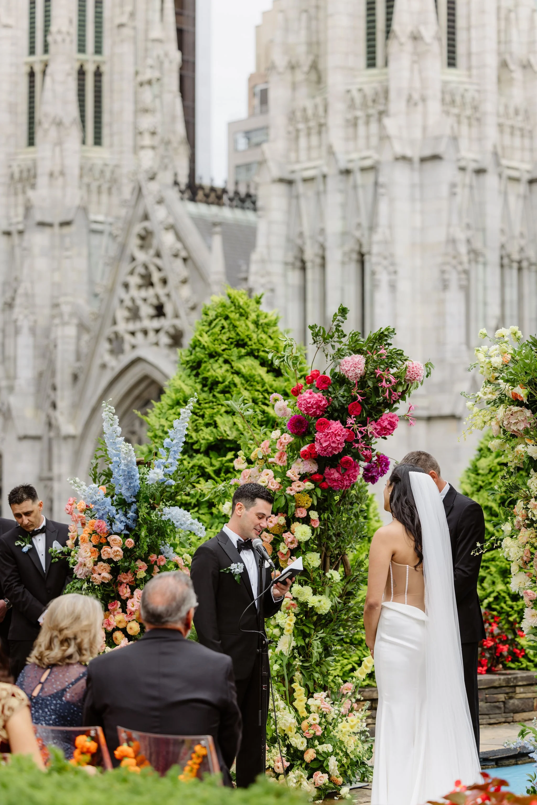 A wedding ceremony taking place outdoors in front of a gothic-style cathedral, with a groom and bride standing before an officiant, surrounded by colorful flowers and guests.