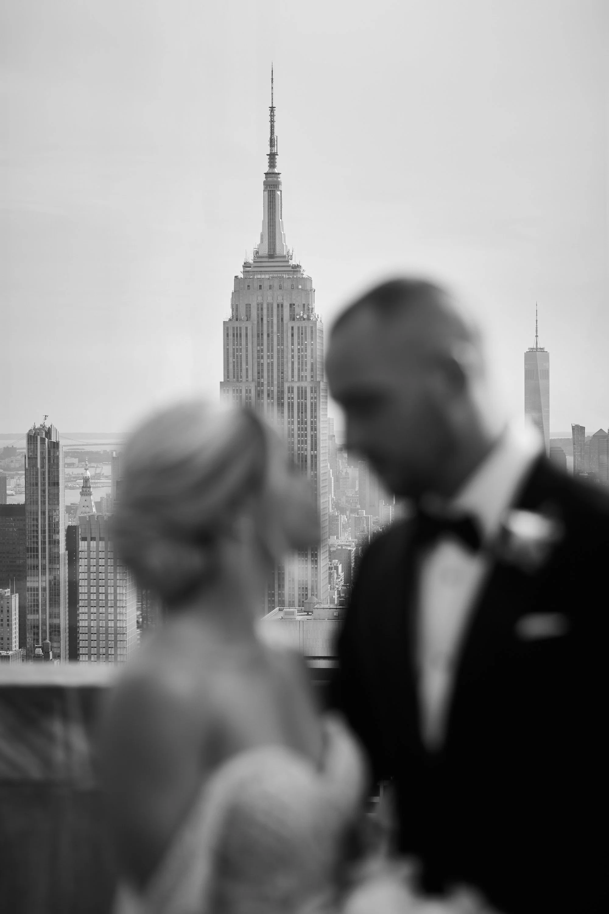 A black and white photo of two blurred figures in formal attire, with a cityscape background featuring the Empire State Building and One World Trade Center.