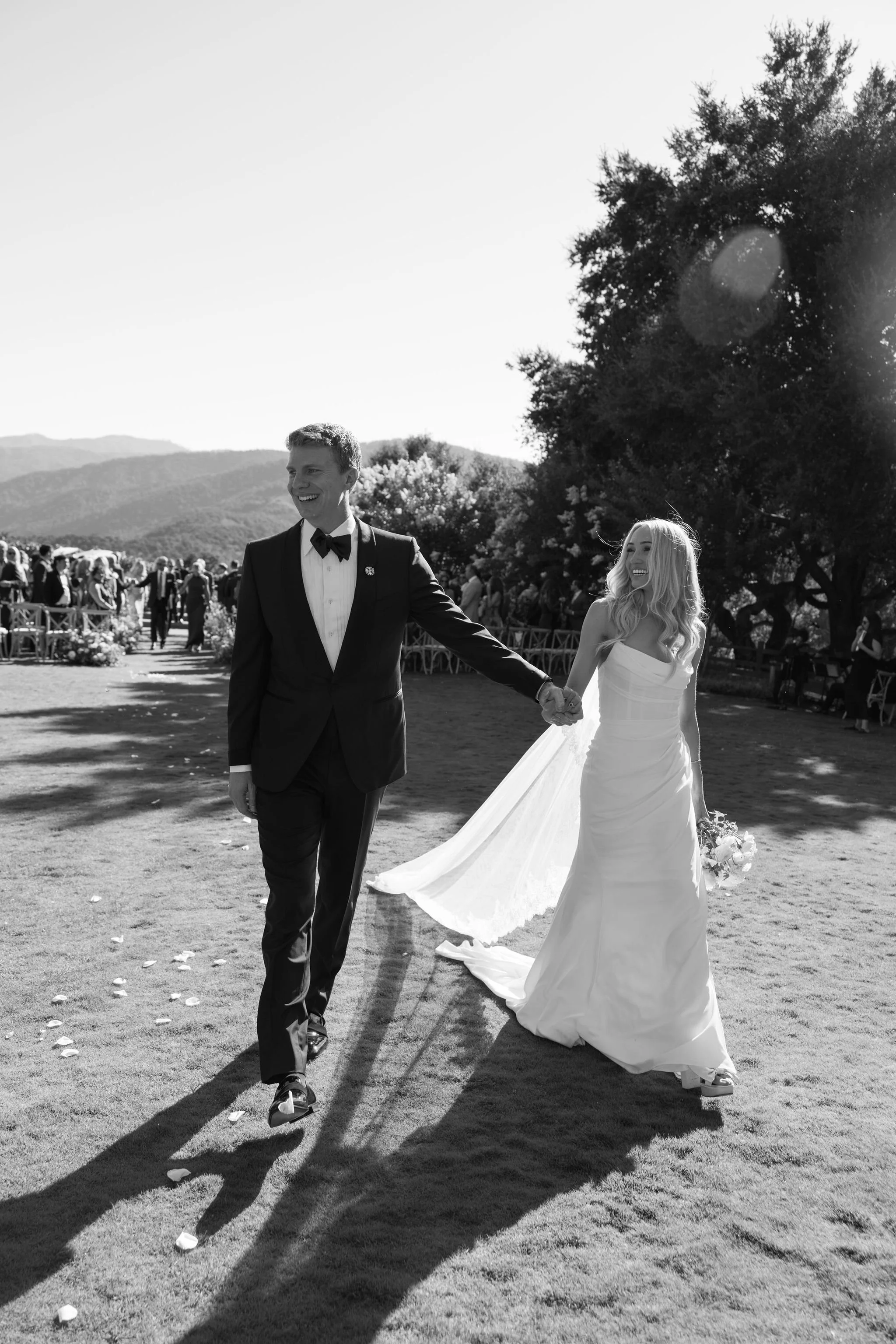 A black-and-white photo of a smiling groom and bride holding hands and walking on a grassy area during a wedding ceremony. The groom is dressed in a tuxedo with a bow tie, and the bride is in a white wedding gown with a long train. There are guests seated in chairs in the background, mountains in the distance, and large trees on one side of the scene.