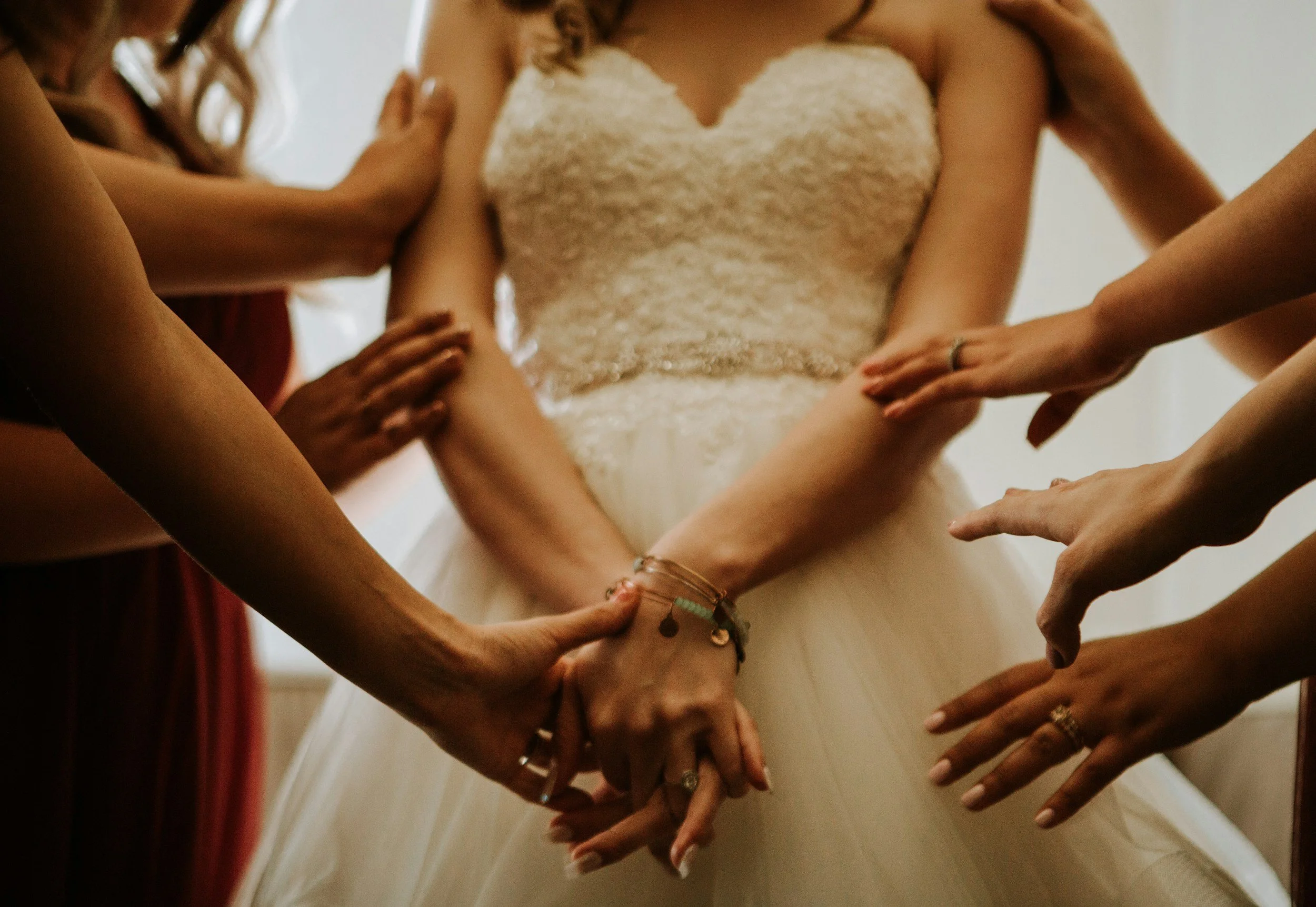 Multiple hands reaching towards a bride, who is wearing a lace wedding dress, touching or adjusting her dress.