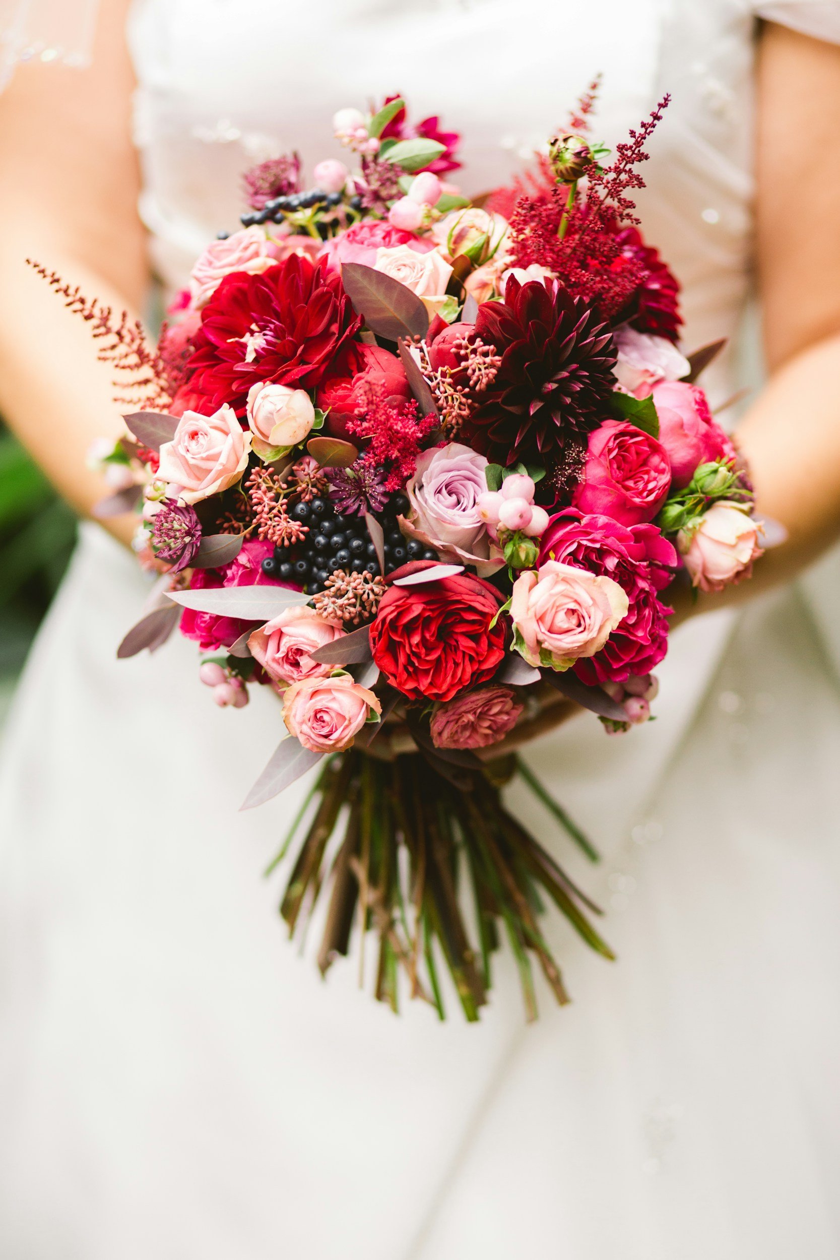 A bridal bouquet featuring pink, red, and purple flowers, with berries and green leaves, held by a bride in a white wedding dress.