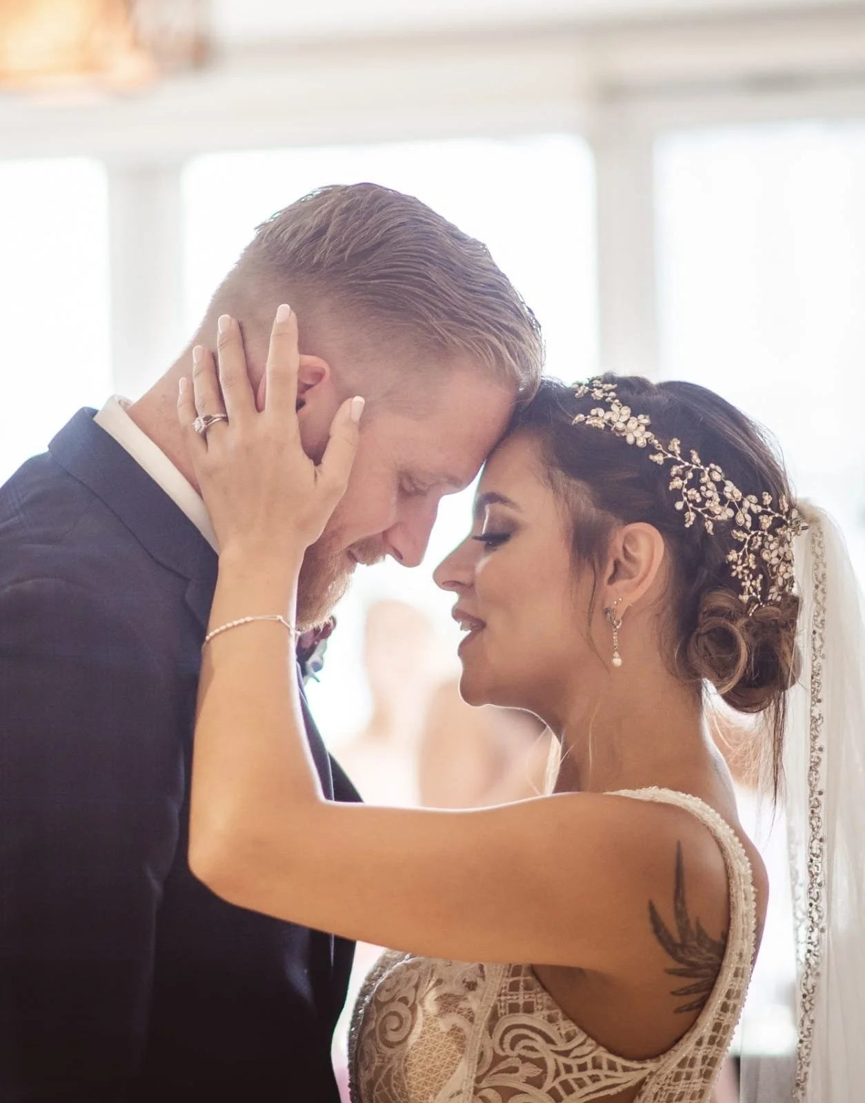 A bride and groom share a close, intimate moment during their wedding, with their foreheads touching and eyes closed. The bride has dark hair styled in an elegant updo with a decorative headpiece, and the groom has short blond hair and is wearing a black suit with a white shirt.