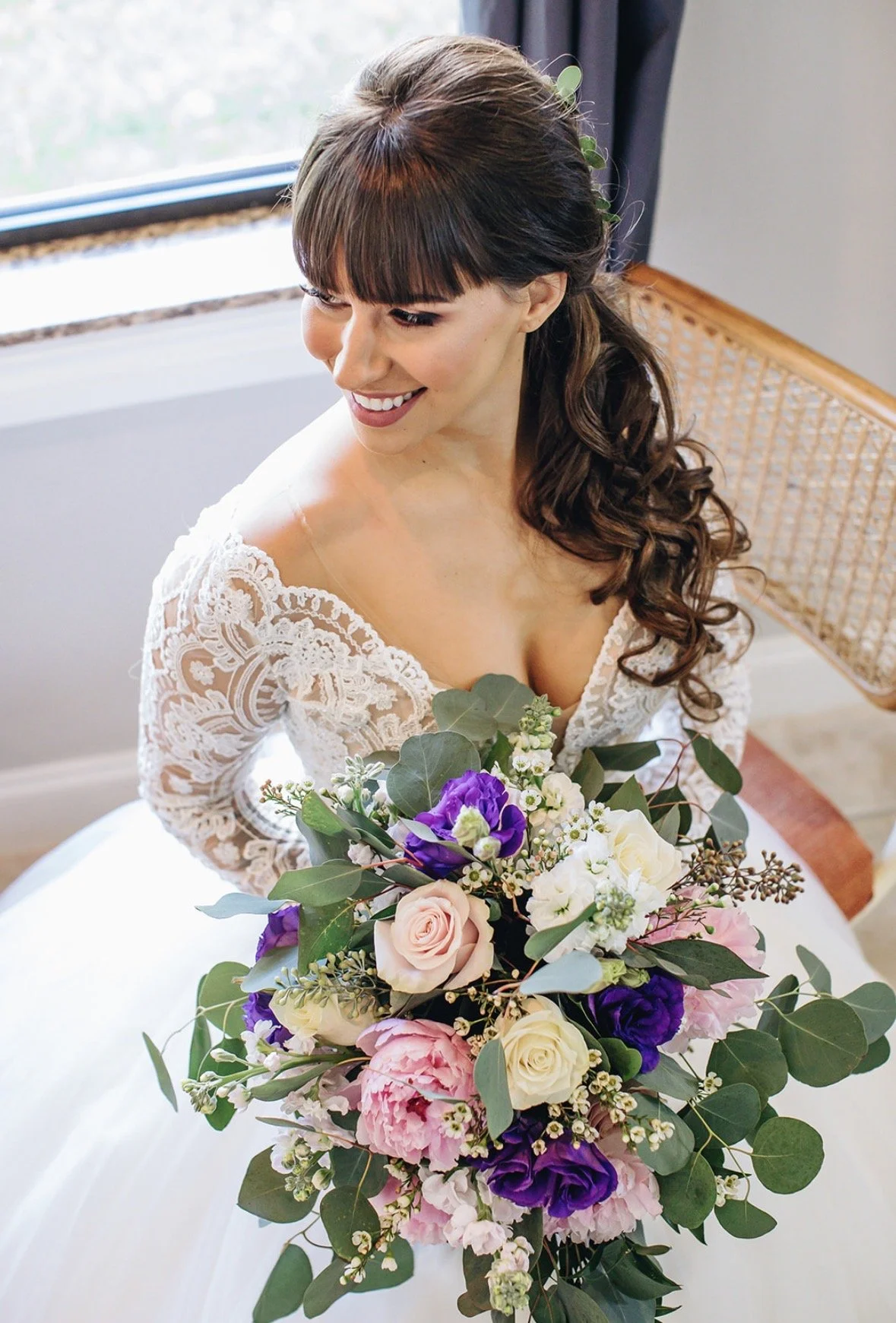 Bride in lace dress holding a colorful bouquet, smiling and looking down, sitting by a window with blue curtains.