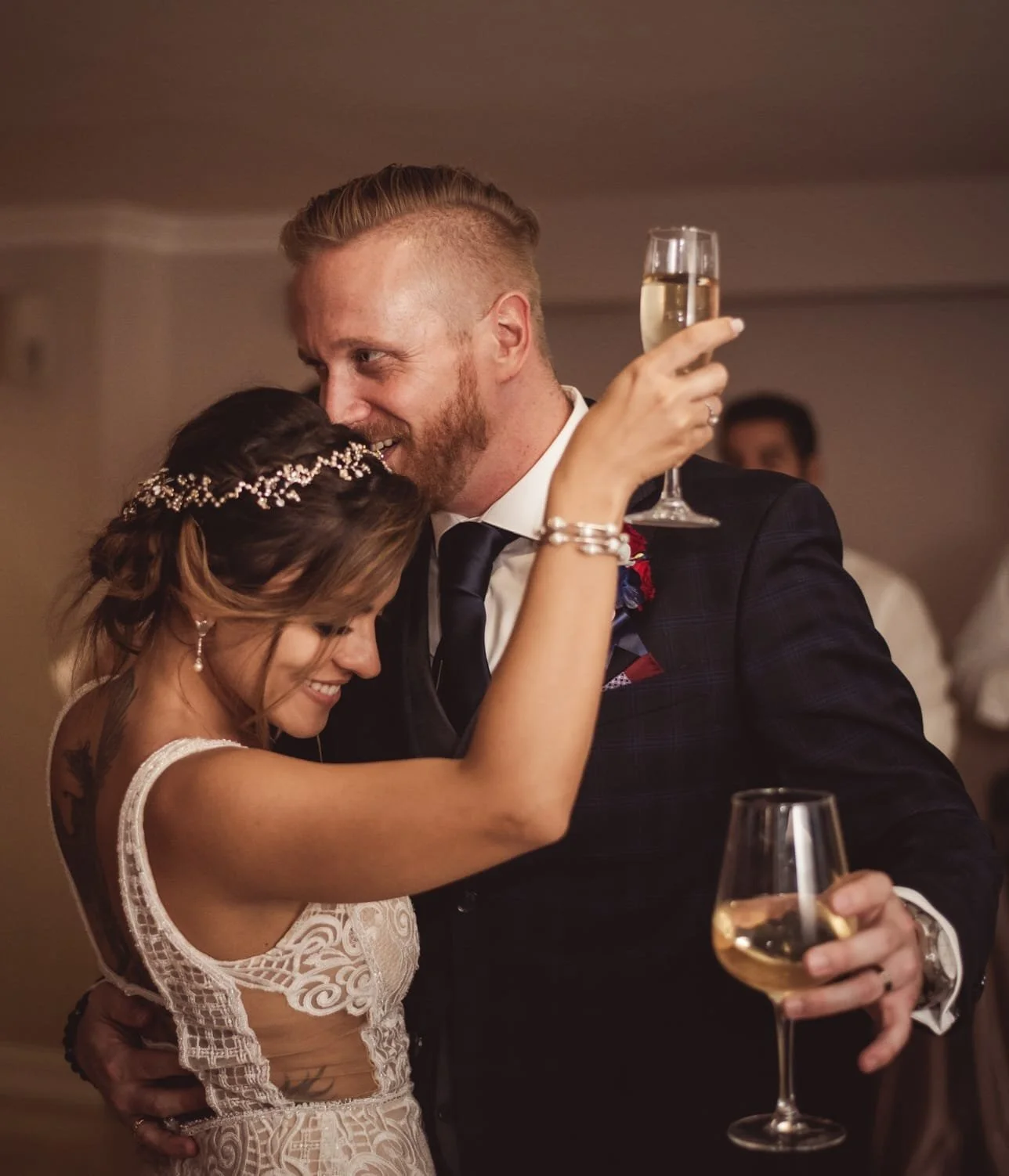 A couple celebrating at a wedding reception, the woman in a white lace dress with a jeweled headpiece, and the man in a dark suit, holding champagne glasses and smiling warmly.