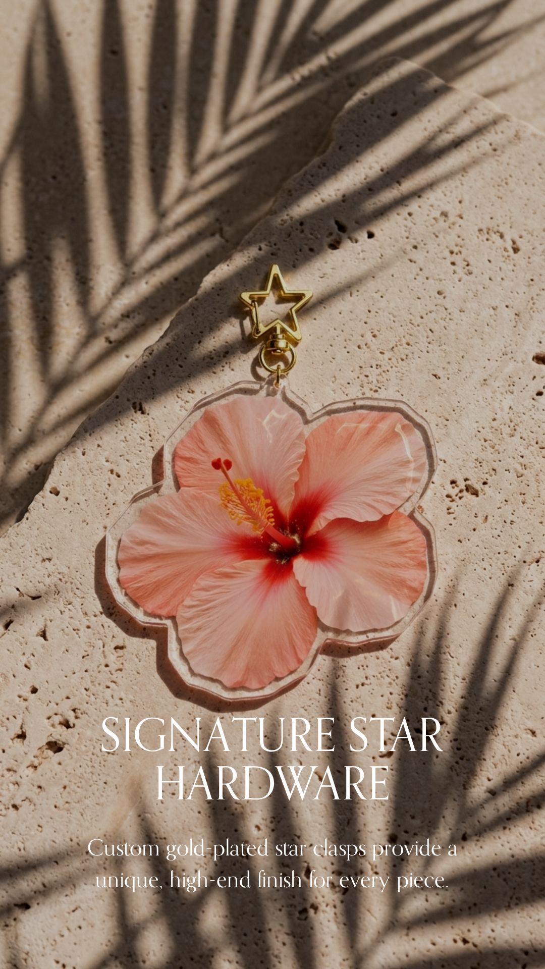 A pink hibiscus flower-shaped keychain with a gold star clasp, placed on a sandy surface with shadows of palm leaves.
