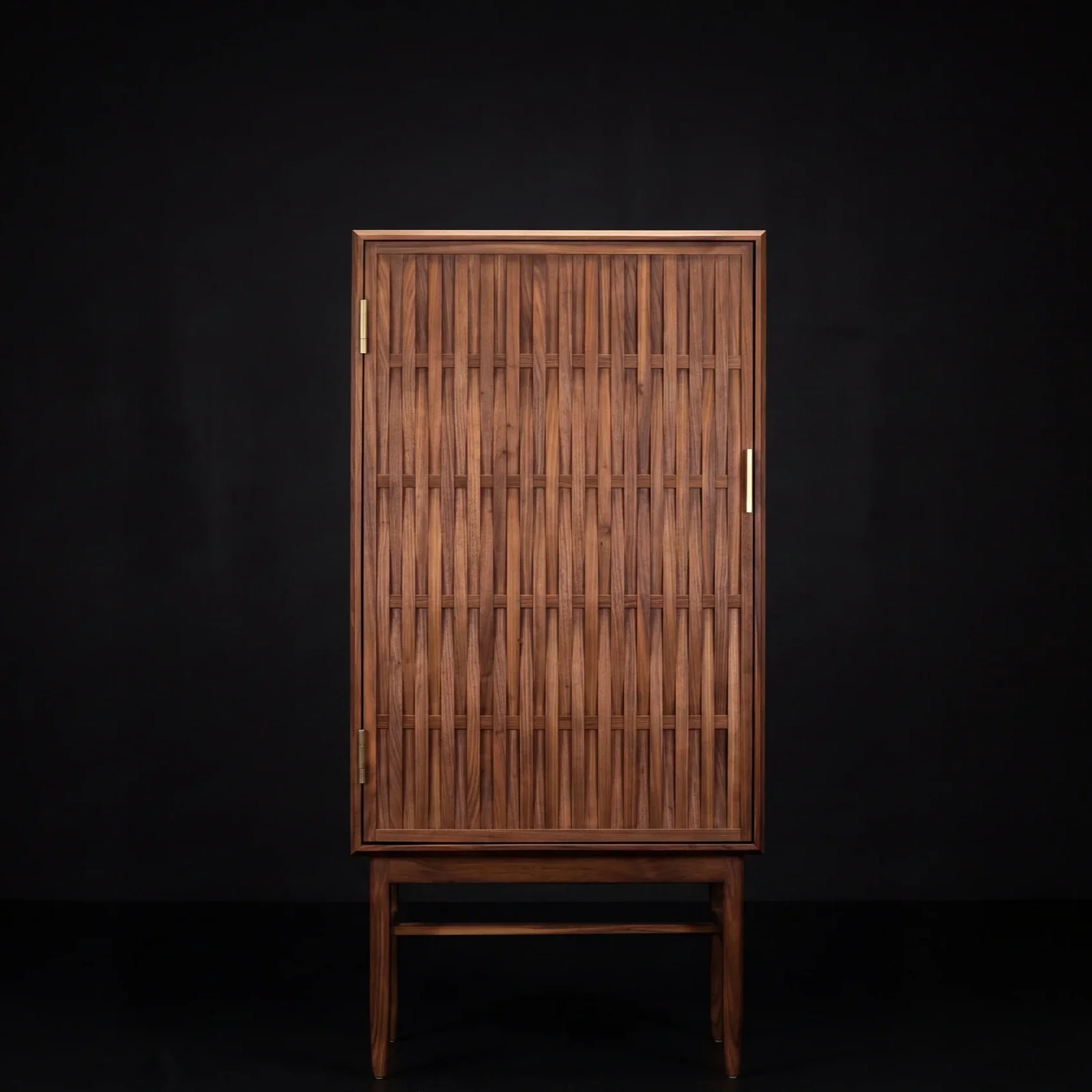 A tall wooden cabinet with a slatted door and brass hinges, set against a black background, with a matching wooden stand at the bottom.