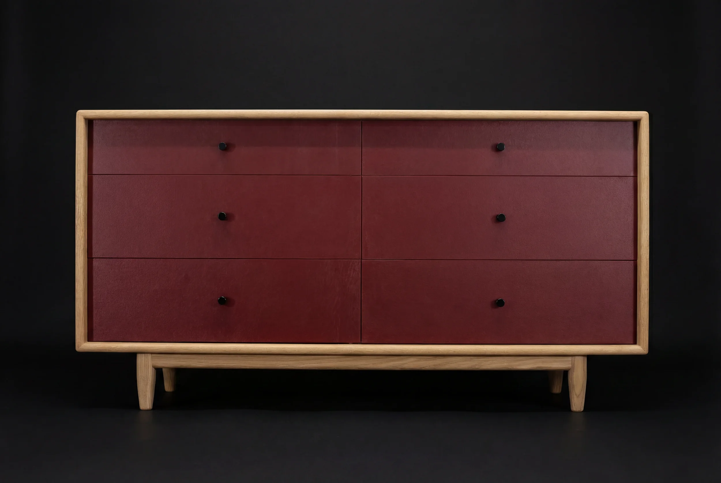 A mid-century modern wooden dresser with six red drawers and black knobs, set against a black background.