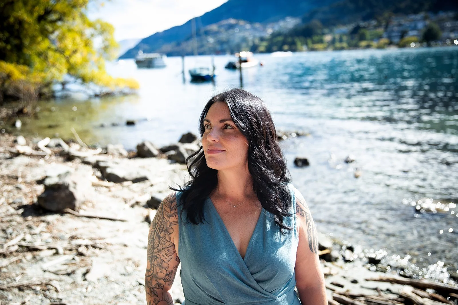 A woman with dark hair, wearing a blue dress, standing on a rocky shoreline by a lake with boats and hills in the background.