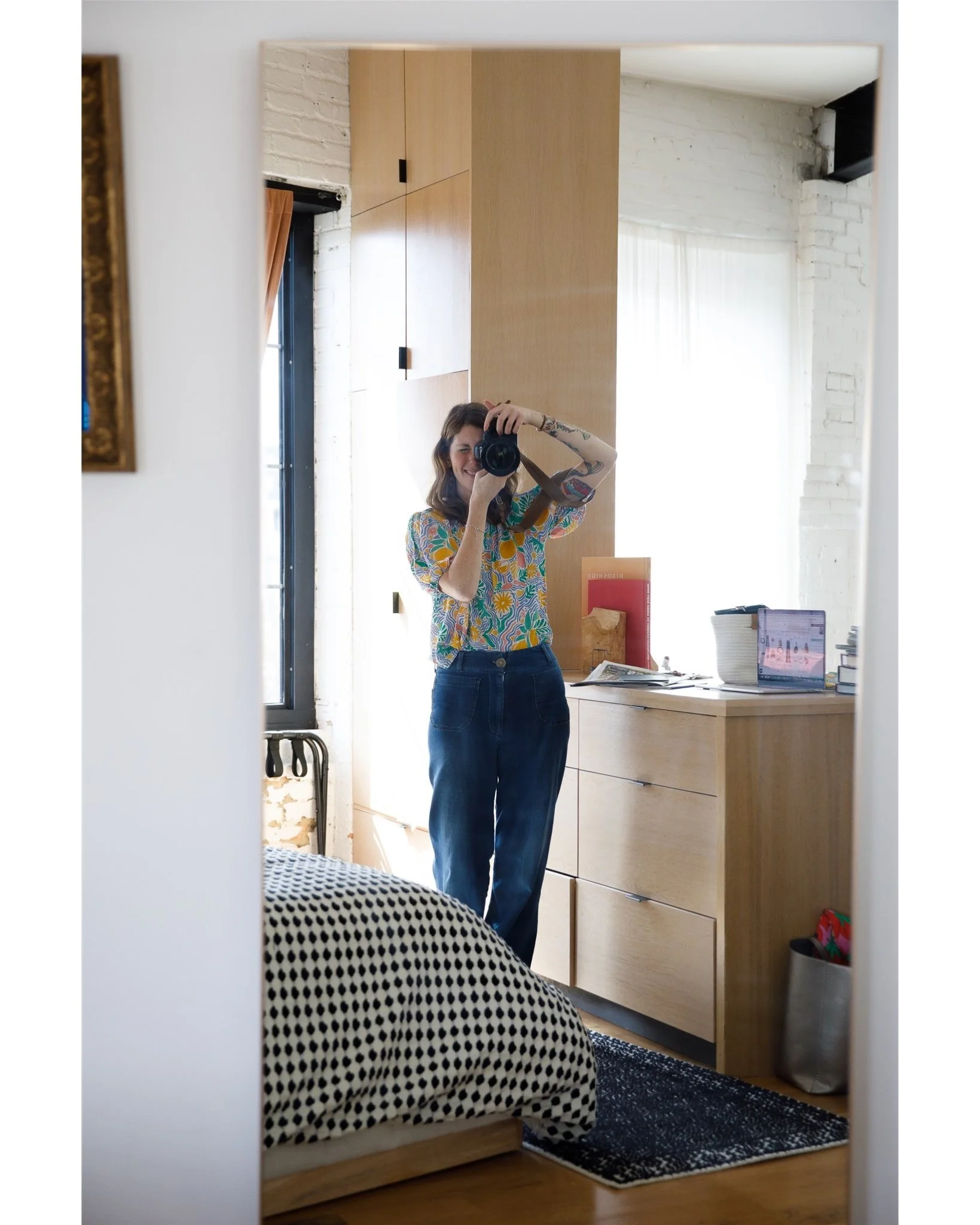 A woman taking a mirror selfie in a room with natural light, wearing a colorful patterned blouse and dark jeans, with a wooden cabinet, laptop, and various objects on a desk visible in the background.