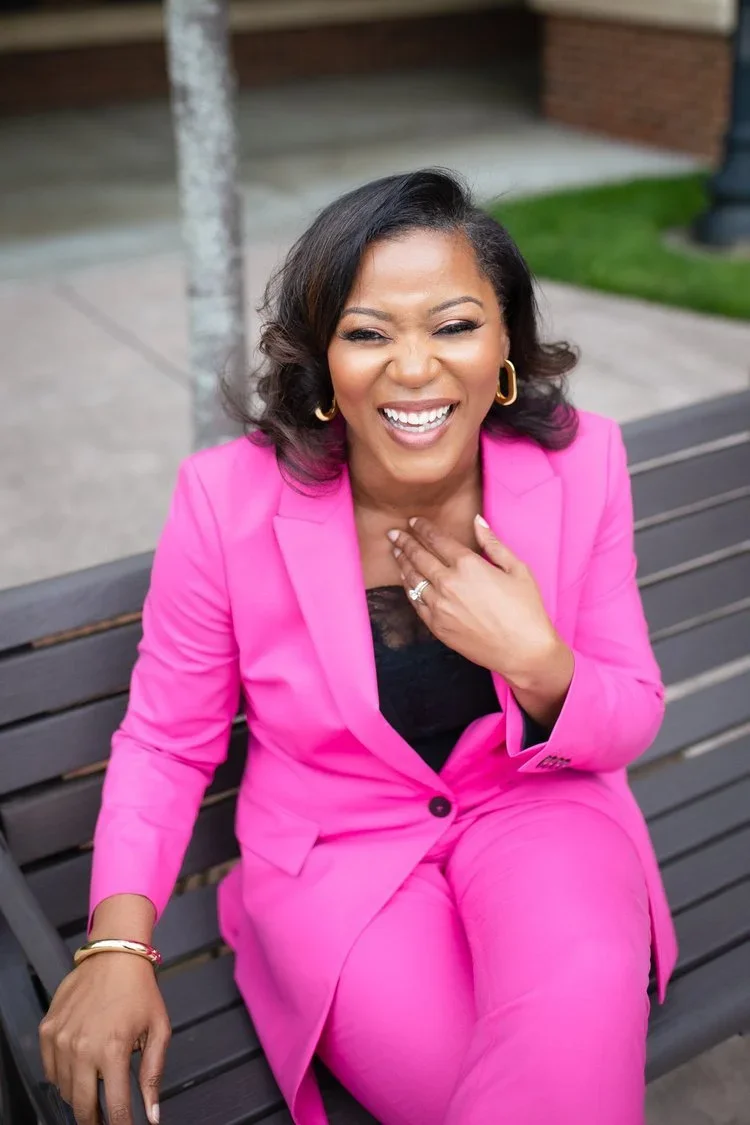 A woman with dark hair wearing a pink suit and gold jewelry sitting on a park bench, laughing and touching her chest.
