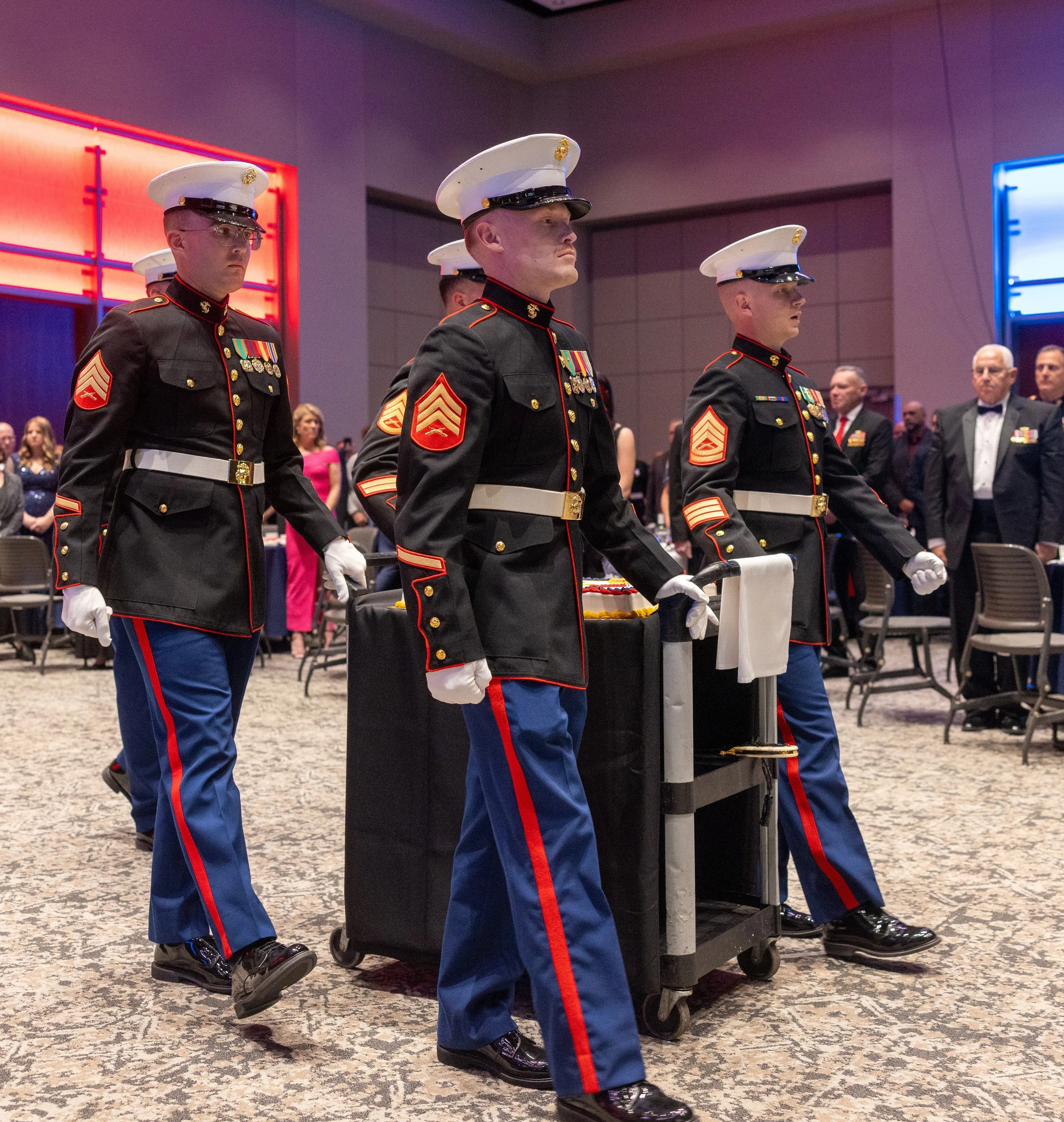 A group of U.S. Marines in dress uniforms participating in a formal ceremony, walking in formation with medals on their chests, in a decorated indoor venue.