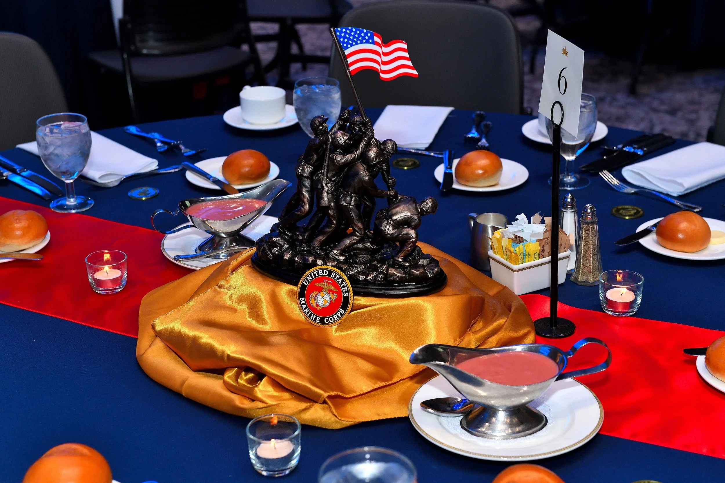 Table decorated for a celebration with a bronze statue of soldiers, an American flag, table number six, candles, glasses, bread rolls, and condiments.