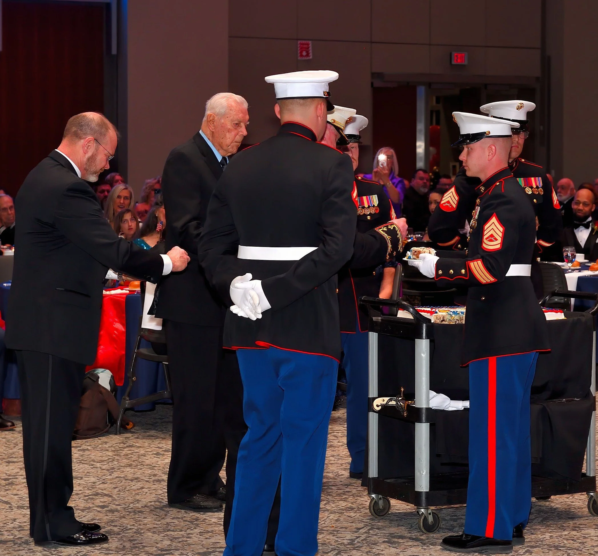 A group of U.S. Marine Corps soldiers in dress uniforms standing in formation at a formal event, with some saluting and others preparing for a ceremony.