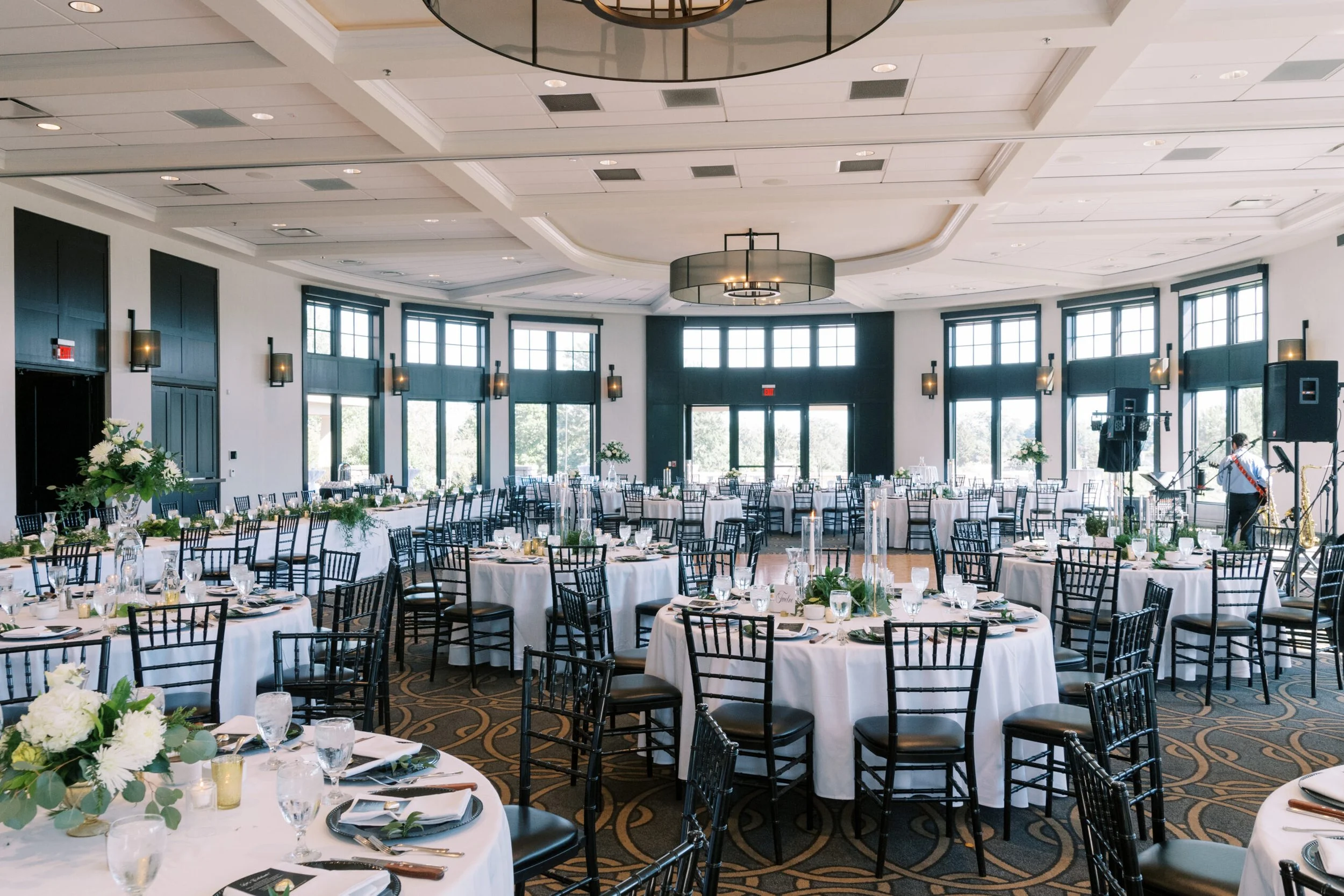 Elegant banquet hall decorated for a formal event with round tables covered in white tablecloths, set with glassware, plates, and cutlery, adorned with floral centerpieces, surrounded by black chairs, with large windows and natural light, and modern light fixtures on the ceiling.