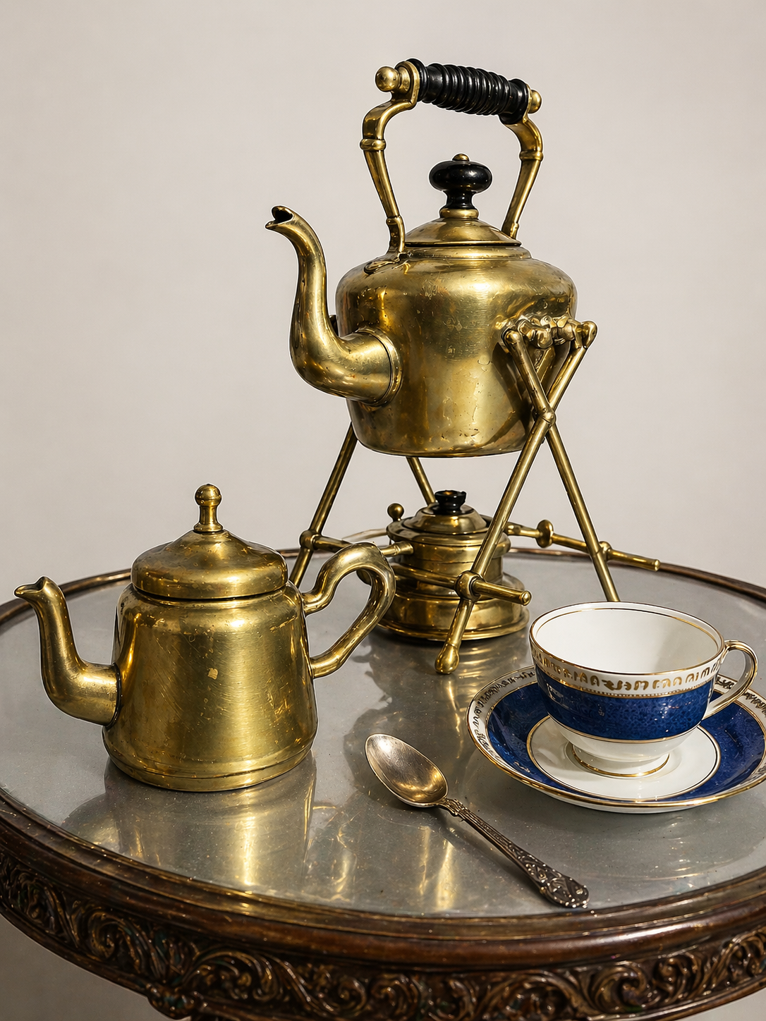 Vintage brass tea set on a round ornate table, including a teapot, a kettle, a small container, and a blue and white teacup with a matching saucer, with a silver spoon beside it.