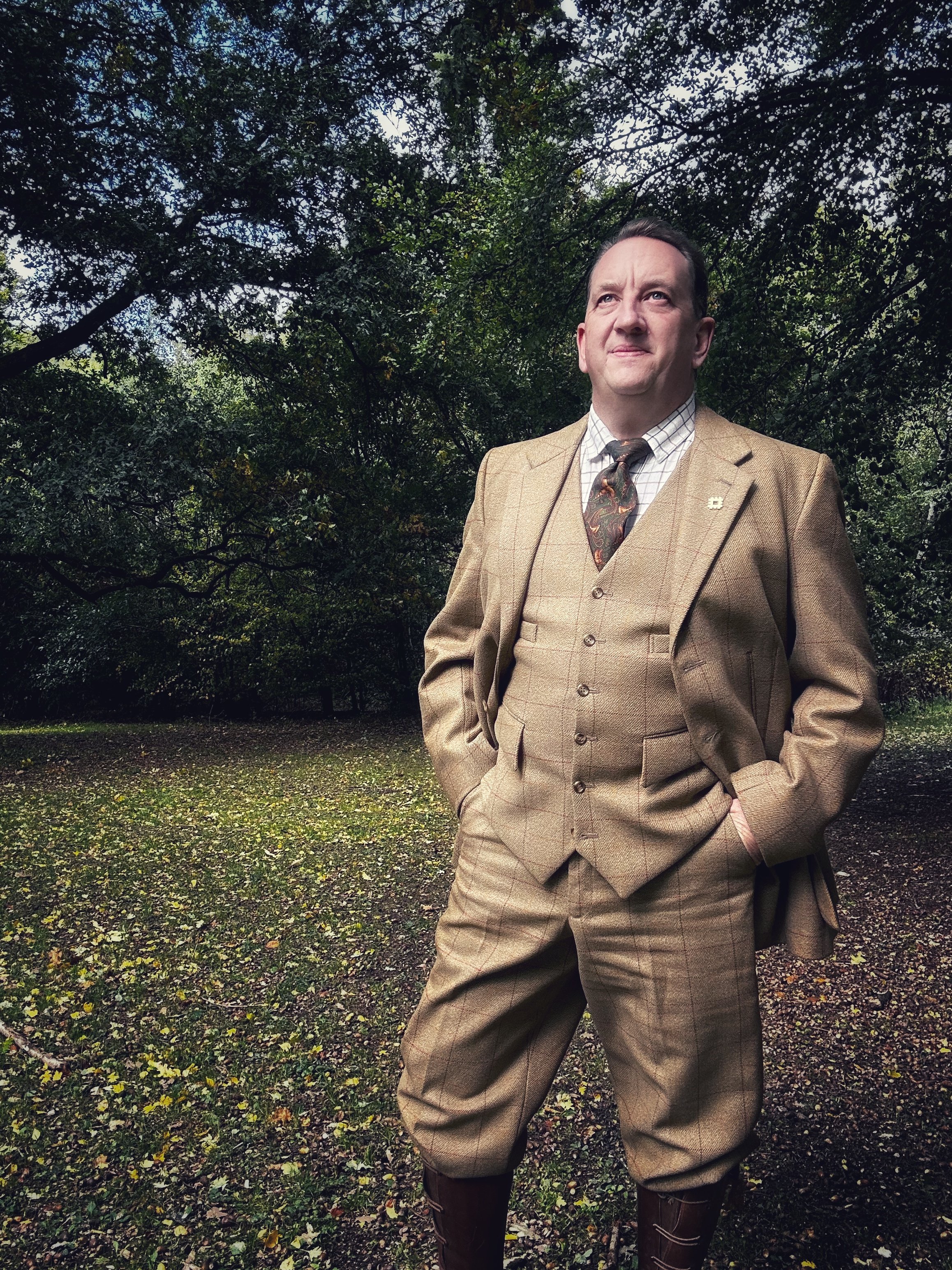 A man standing outdoors in a wooded area, wearing a tan plaid suit with a white checked shirt and a patterned tie, with his hands in his pockets and a confident expression.