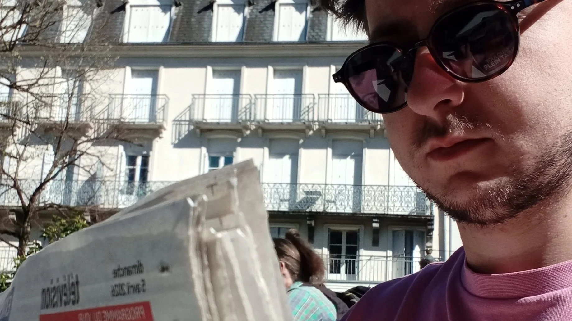 Un homme portant des lunettes de soleil regarde un objet dans ses mains, avec une façade d'immeuble en arrière-plan.