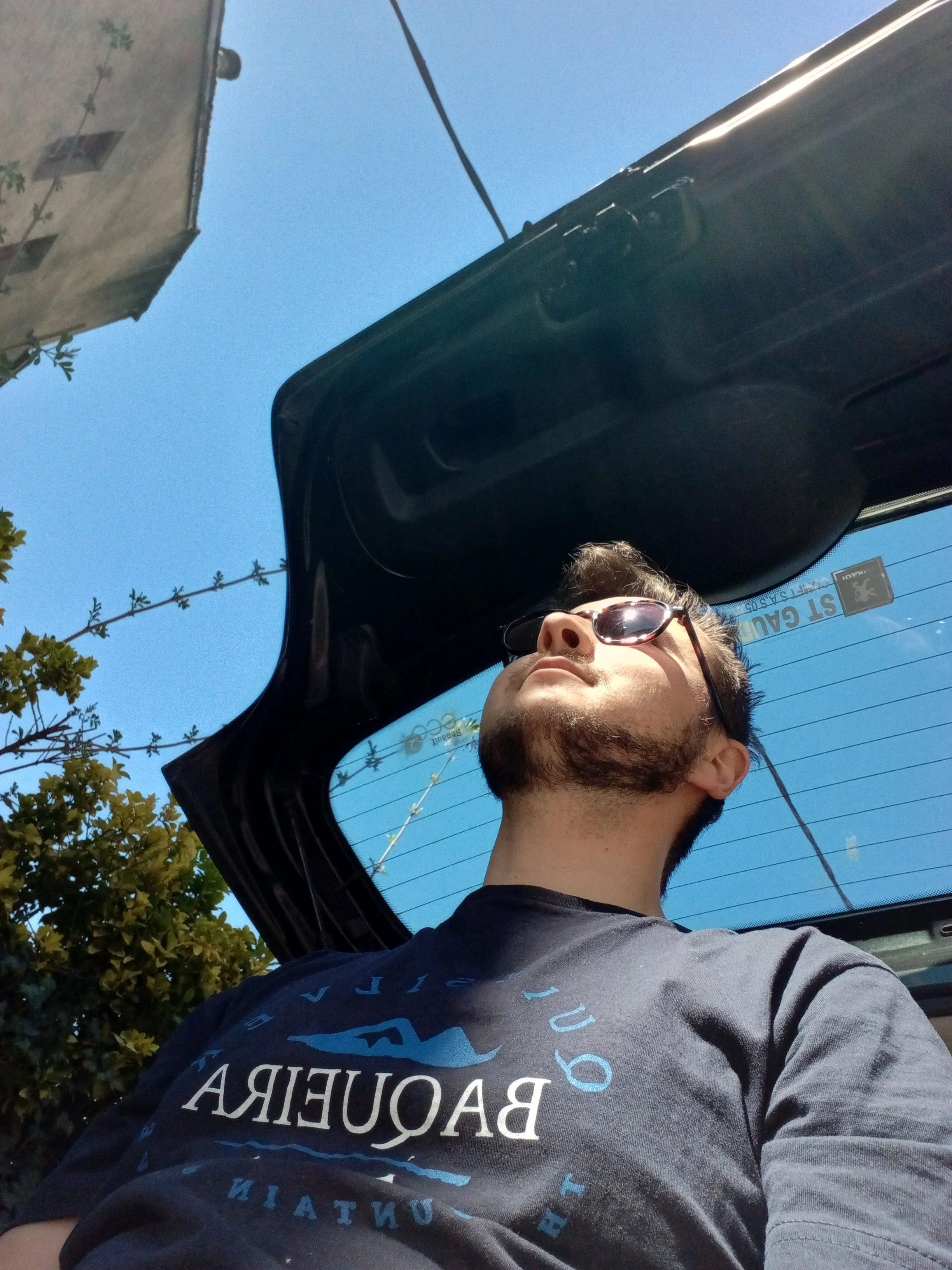 Une personne portant des lunettes de soleil et un t-shirt gris clair avec des écritures bleu clair, vue de profil, dans une voiture ouverte avec un ciel bleu et des arbres en arrière-plan.