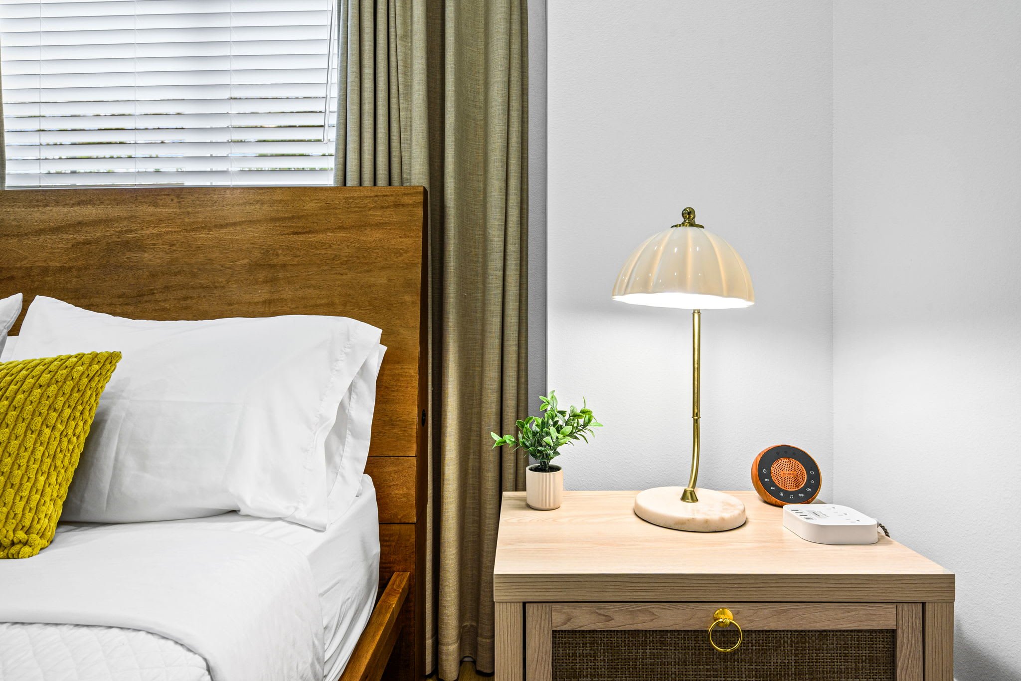 A bedroom with a wooden bed frame, white bedding, and a yellow textured pillow. There is a window with white blinds and beige curtains. Next to the bed, there is a wooden nightstand with a plant, a lamp, an orange and black speaker, and a white electronic device.